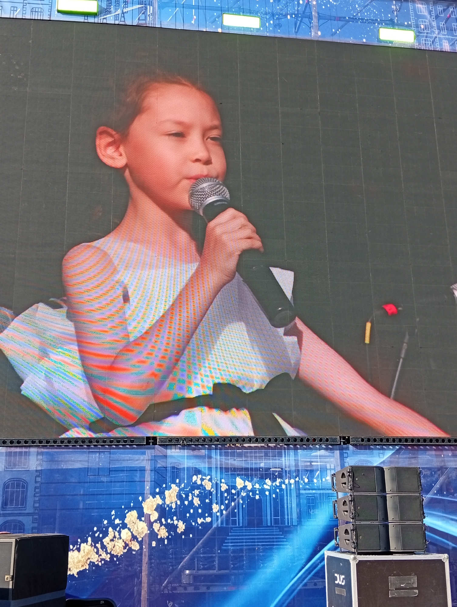 A young girl, with her hair tied back and her eyes looking to her left, stands on a stage, holding a microphone to her lips. She is wearing a white off-the-shoulder dress. The background is a large LED screen displaying a green grid pattern with a colorful, abstract image of a building at the top. Below the LED screen, another screen displays a blue background with white, abstract shapes resembling clouds or scattered light. To the right of the lower screen, a stack of black speaker cabinets sits on top of a black case. In the foreground, a black speaker box is visible on the left. The overall scene appears to be an outdoor performance or event. The location is the city of Kostanay, Kazakhstan.