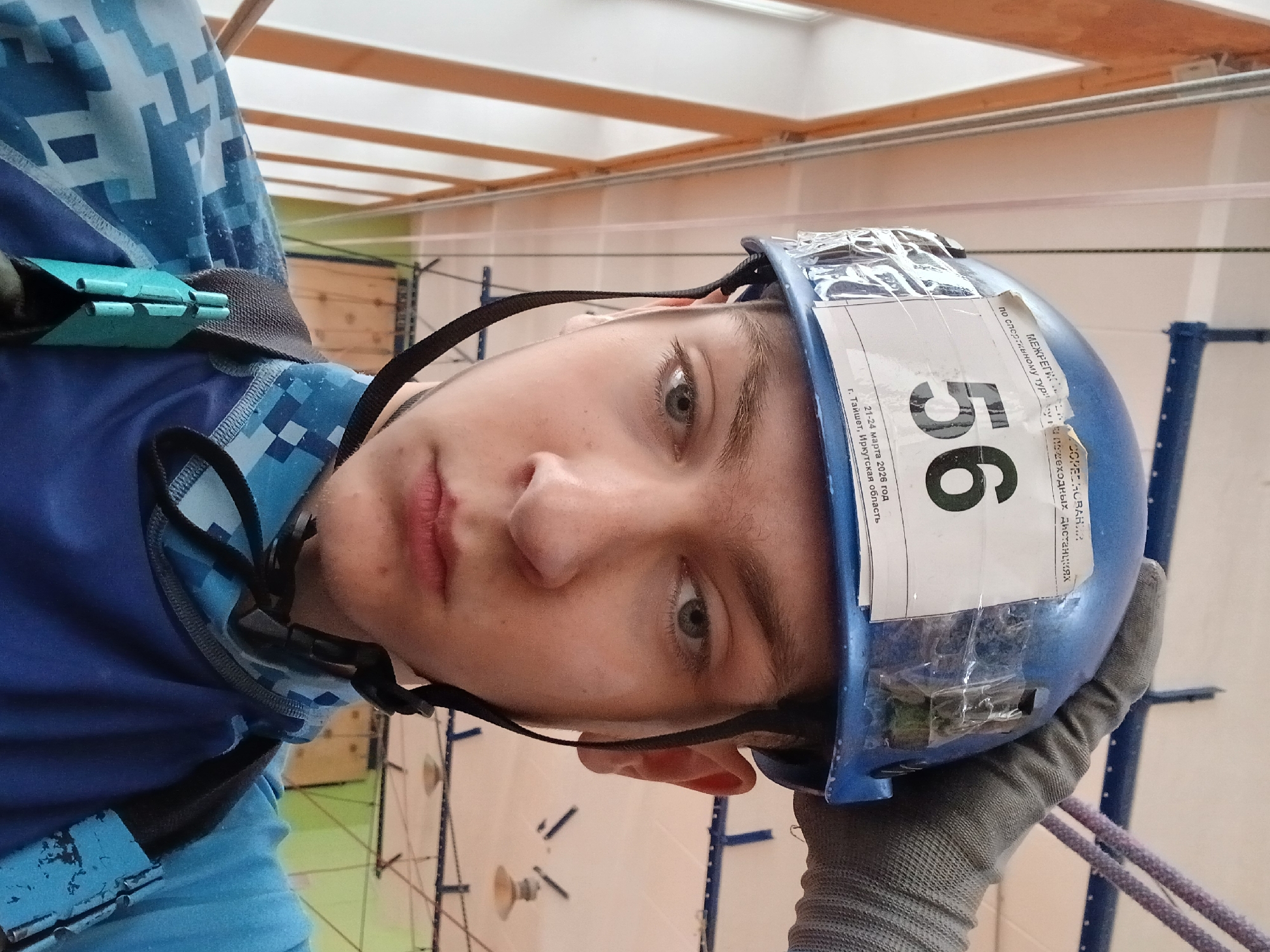 A young male wearing a blue climbing helmet with the number "56" displayed on a white sticker is positioned indoors, likely in a climbing facility. He's also wearing a blue shirt with a pixelated pattern and a climbing harness. He has one gloved hand visible. The image is rotated, showing the person's face angled towards the camera. In the background, there's a wooden structure with beams, climbing equipment, and white walls. The text on the sticker mentions "21-24 марта 2026 год" and "г. Тайшет, Иркутская область". There is no indication that the location is Chaikovsky.