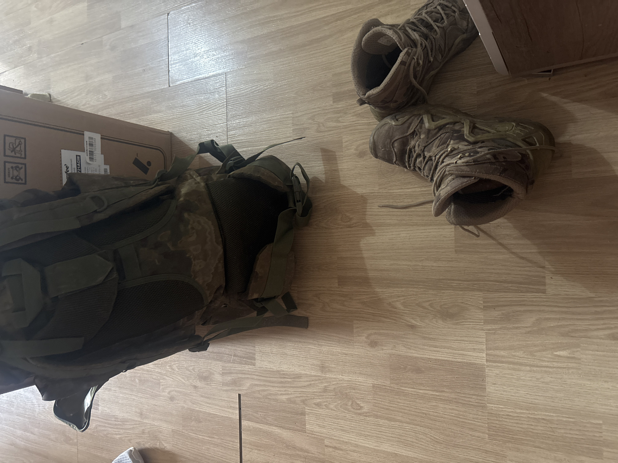 The image shows a room with a light-colored, wood-patterned floor. A military-style backpack sits on the left side, partially obscured by a cardboard box. To the right, a pair of tan, high-top hiking boots rest on the floor. The location and presence of people cannot be determined.