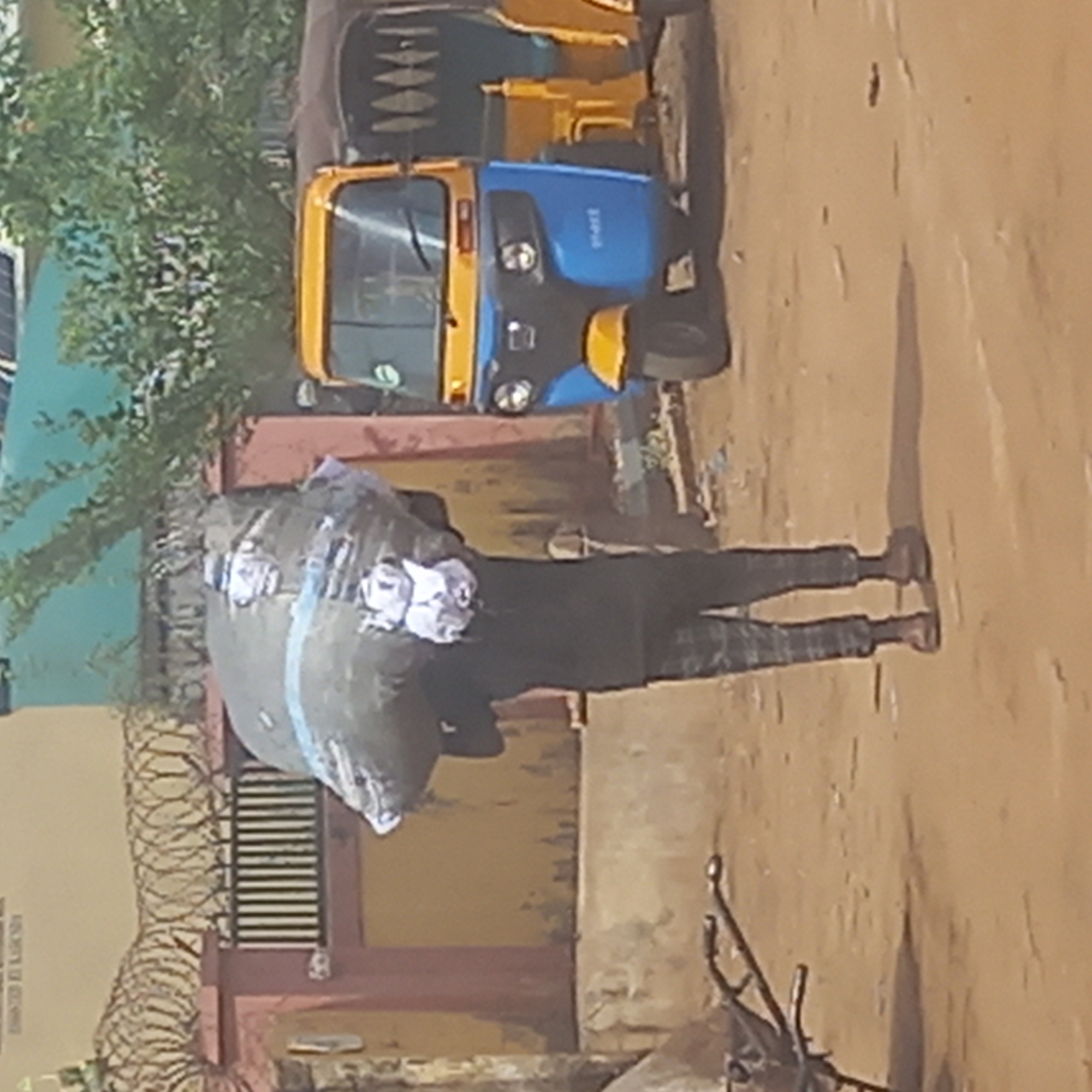 A man carries a large, wrapped bundle on his head in Dadi Riba, Nigeria. He is walking on a dirt road next to a blue and yellow commercial tricycle. In the background, a building with barbed wire on top of the wall is visible. There are also some trees in the background. The scene appears to be a street in the city.