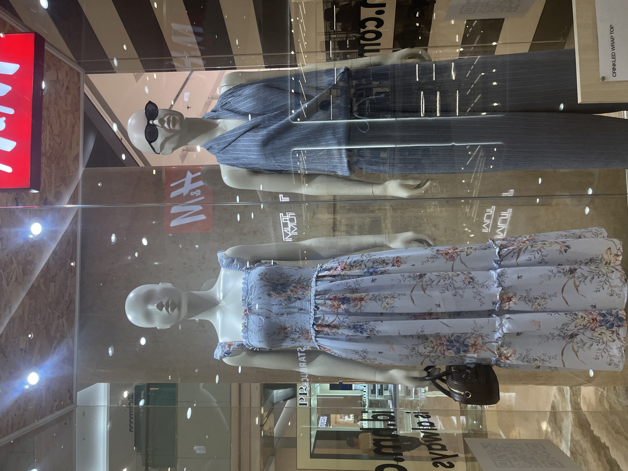 A store window display features two mannequins. The mannequin on the left wears a light blue floral print midi dress with ruffled straps and hem. It carries a small brown handbag. The mannequin on the right wears a grey pleated wrap top and matching wide-leg pants. It has dark sunglasses on. Behind the mannequins, a red H&M logo is visible, along with reflections of the surrounding mall interior. The scene is located in South Jakarta, Indonesia.