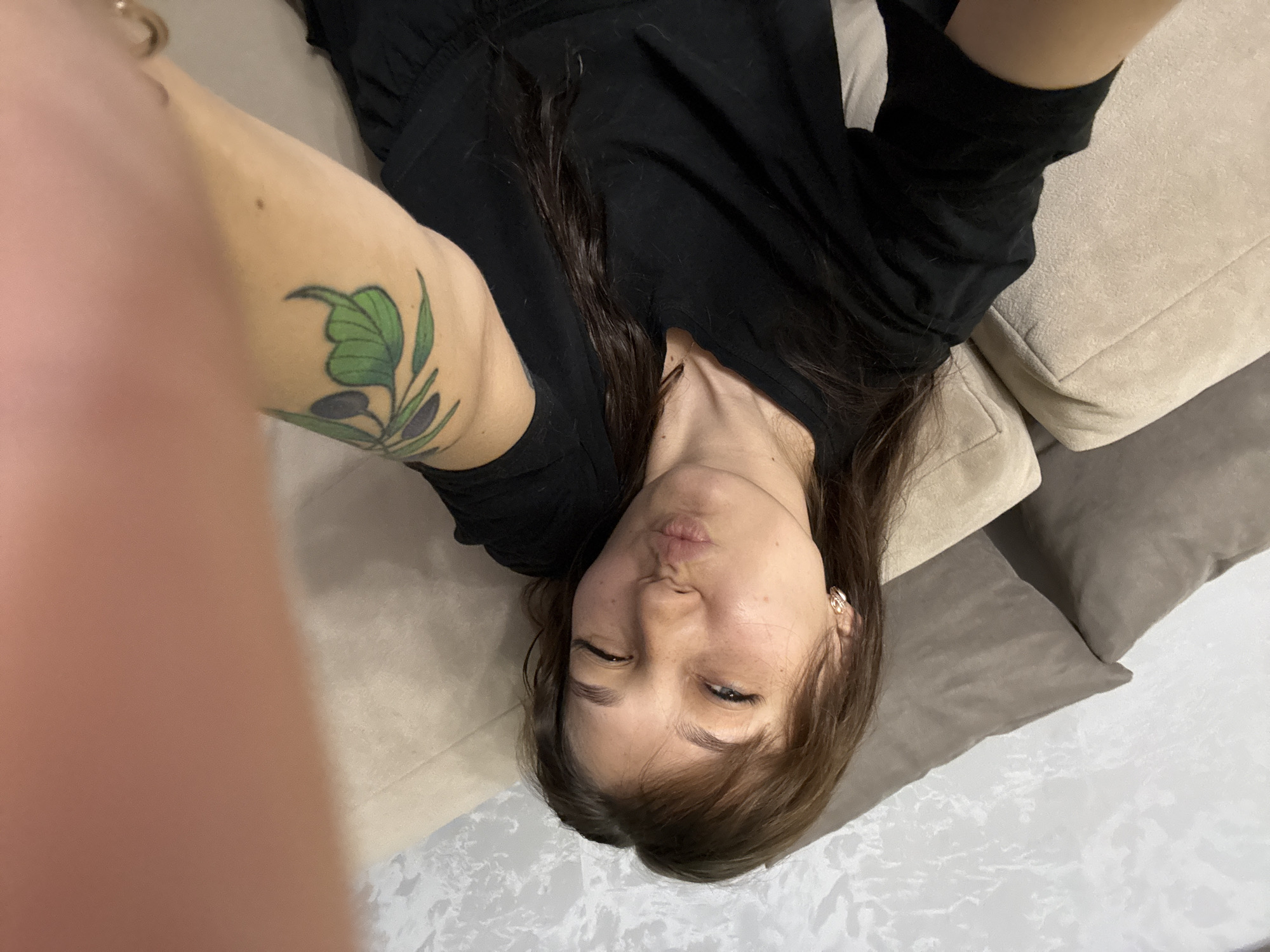 A young woman with brown hair is taking a selfie on a couch. She is wearing a black t-shirt and has a tattoo of an olive branch on her left arm. The background appears to be a living room with a textured wall and plush cushions on the couch. The image was taken indoors.