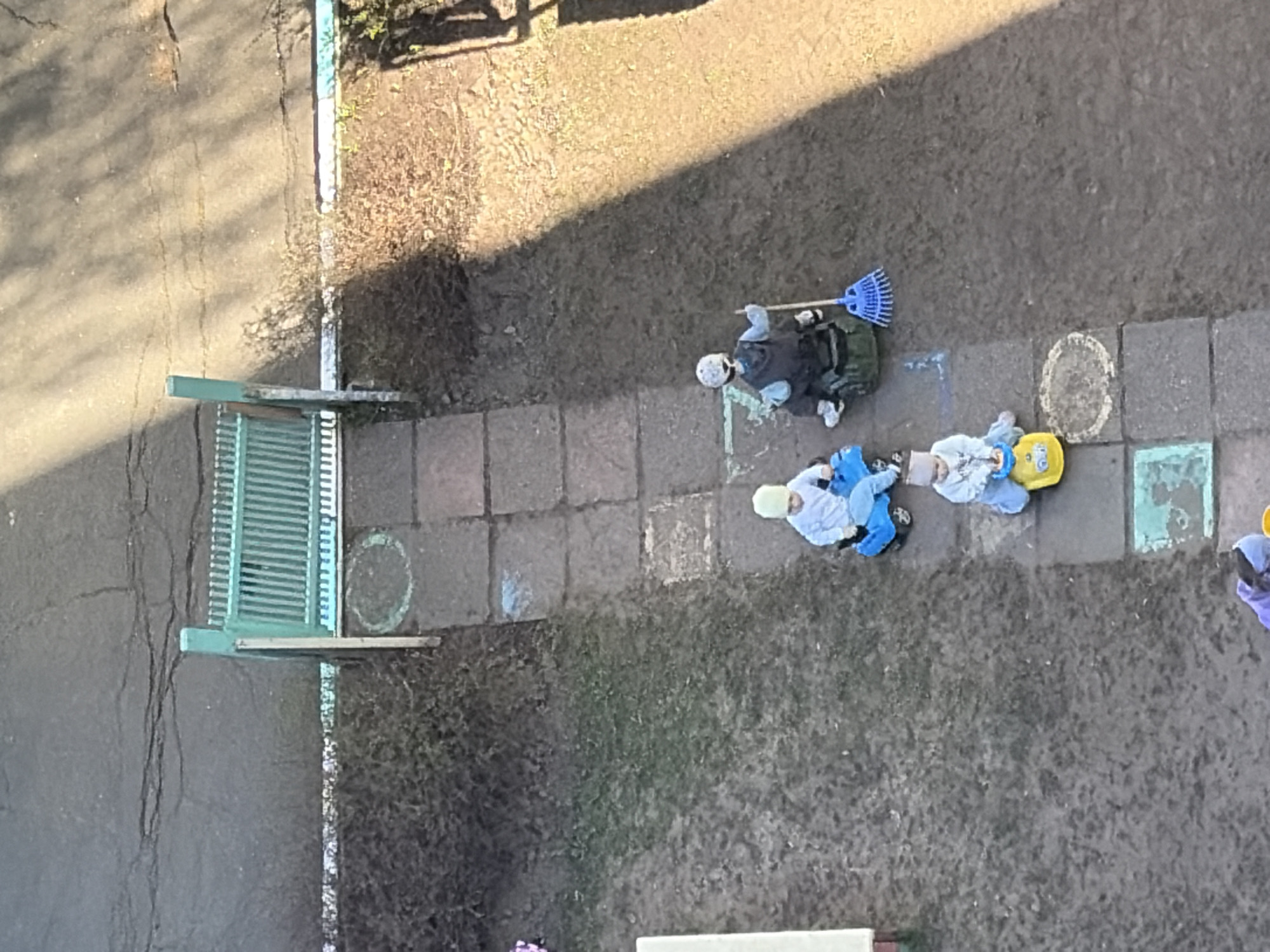 An aerial view depicts a courtyard scene in Izhevsk, Russia, adjacent to a building and featuring a walkway constructed of rectangular pavers. A green park bench is visible on the left side of the frame, near the walkway. Several individuals, possibly children, are present, dressed in outerwear and appearing to be engaged in outdoor play. One individual carries a blue rake. There are circular markings on the pavers, potentially part of a game or activity. The surrounding area consists of a mix of paved surfaces and patches of dirt and grass.