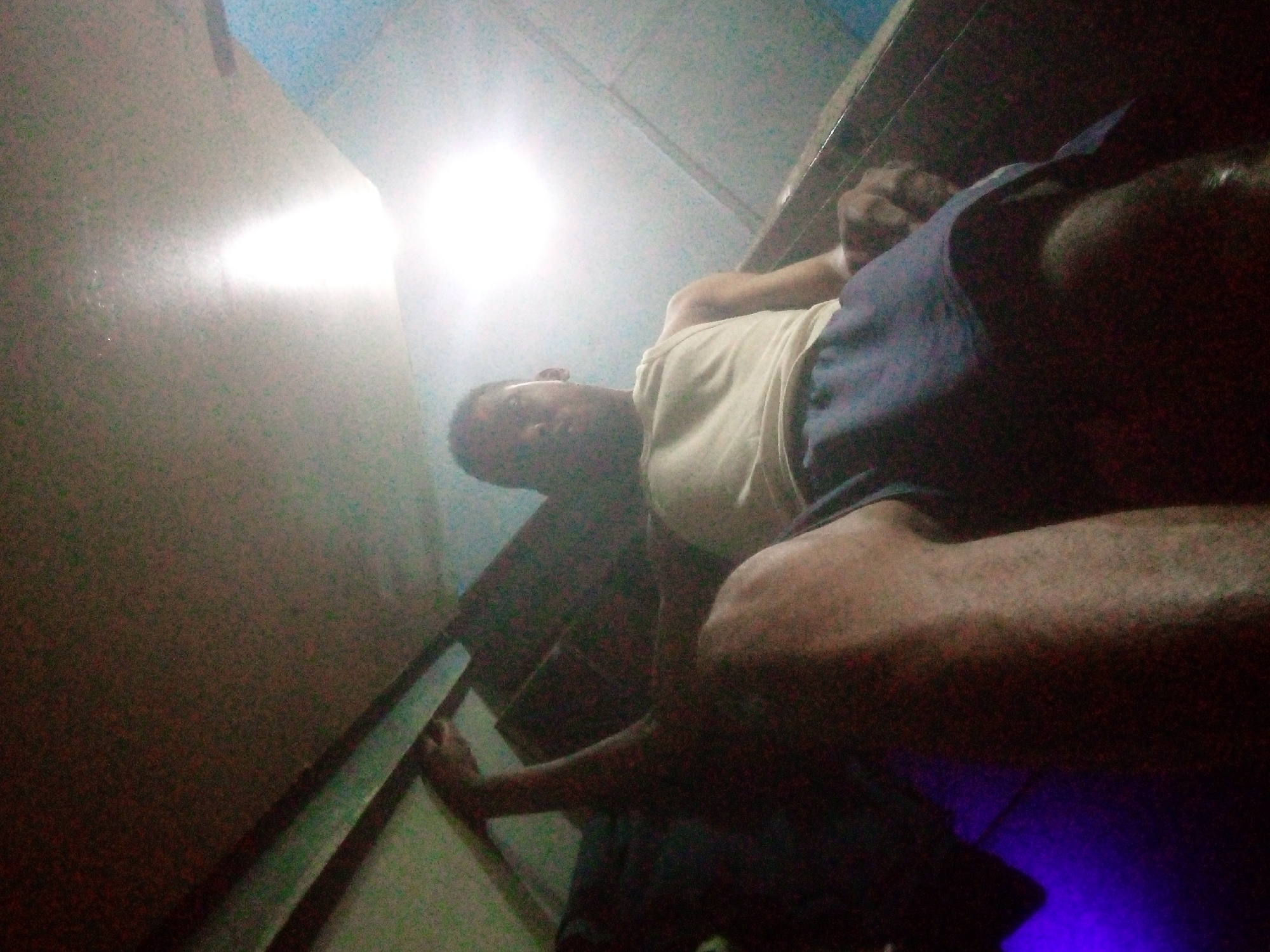 A man wearing a light-colored t-shirt and dark shorts is lying on a bed or couch in a room with a tiled ceiling. The lighting is dim, and a bright light source is visible on the ceiling. A dark-colored wall or object is to the left. The location is unknown.