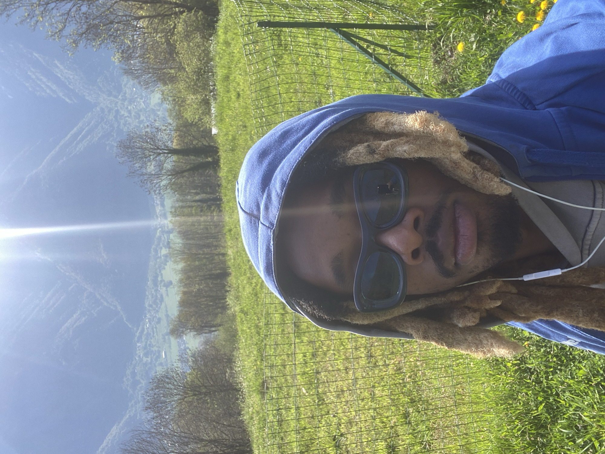 The image, rotated 90 degrees clockwise, features a person with light brown dreadlocks and a beard, wearing a blue hooded sweatshirt and dark sunglasses. A white wired earphone is visible in their right ear, and they are looking directly at the camera. A strong sun flare is prominent on the upper left side of the frame, extending across the person's face and sunglasses.
The setting is outdoors on a bright, sunny day. Behind the person, a green wire mesh fence runs horizontally across the frame. Beyond the fence, the landscape reveals a vibrant green grassy area dotted with yellow dandelions in the mid-ground. Further in the background, a sprawling valley with trees, fields, and distant mountains or hills unfolds under a clear blue sky. Wispy white contrails or clouds are visible high in the sky. The overall scene suggests a scenic rural or mountainous location, identified as Saint-Georges-d'Hurtières, France. There is no visible text in the image.