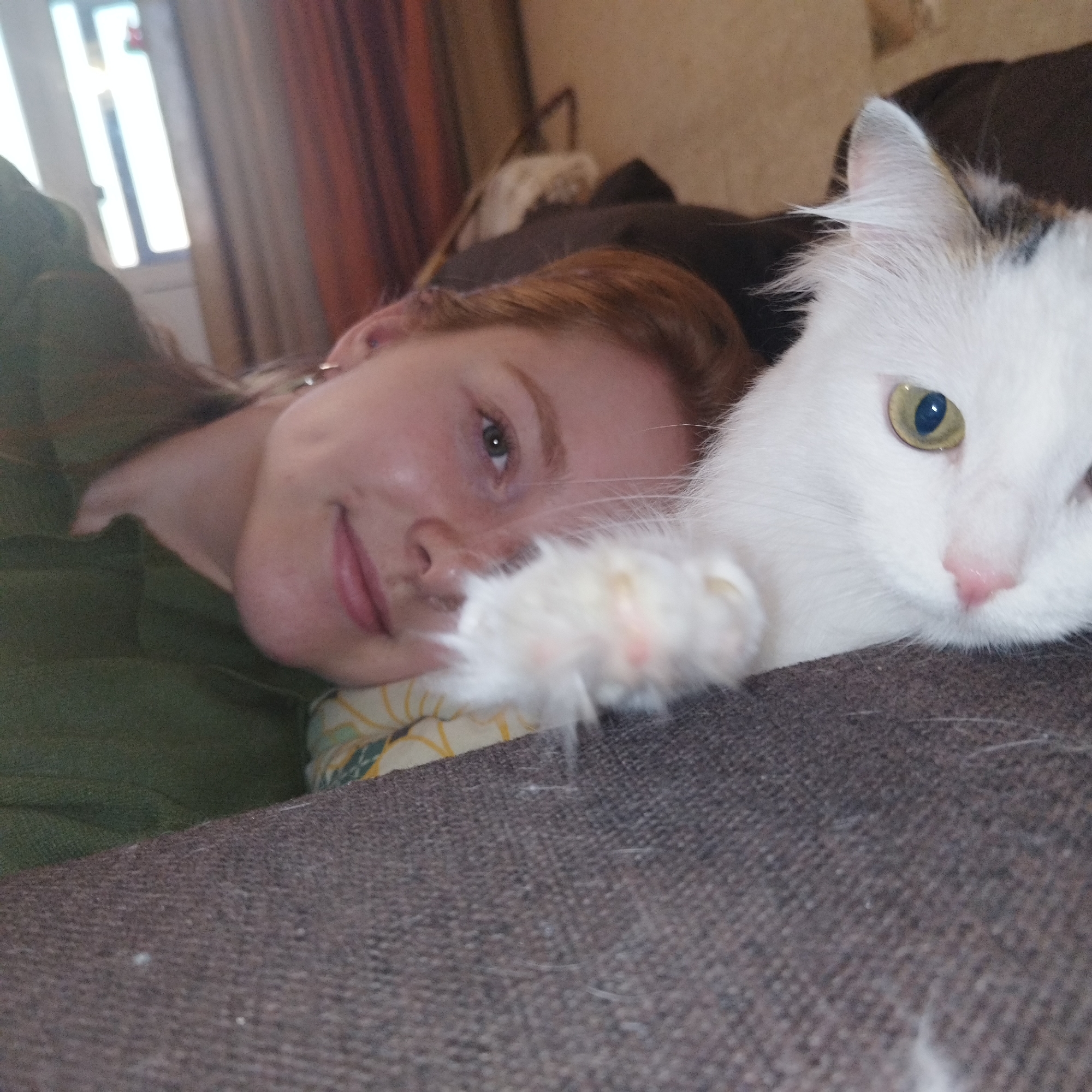 In Yekaterinburg, Russia, an indoor scene features a female subject and a domestic cat resting together. The female, with reddish-brown hair, fair skin, and green eyes, is observed lying on her left side, resting her head on a patterned pillow. She exhibits a subtle smile and is dressed in a dark green garment, with a silver earring present on her left ear. A predominantly white cat, displaying dark markings near its right ear, is positioned adjacent to the female. The cat possesses yellow irises and a pink nose; its left paw, showing pink pads, is extended into the foreground. Both subjects are oriented towards the camera. The foreground consists of a dark grey, textured upholstered surface. The background features a brightly lit window with white vertical framing, adjoined by a rust-colored curtain.