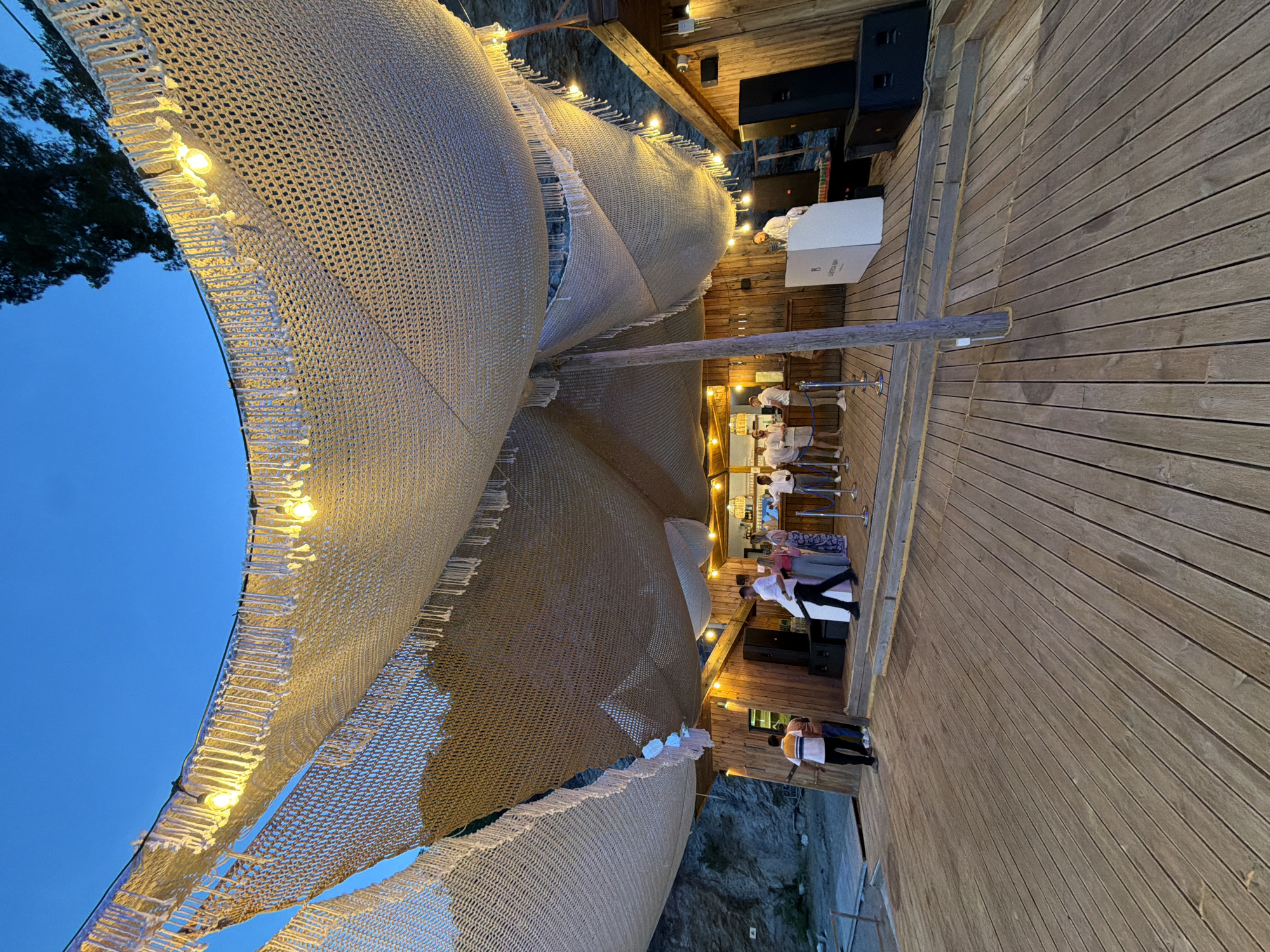 The image shows an outdoor event venue in Glavaticici, Montenegro, at dusk. The venue features a wooden deck with a covered area created by a network of netting illuminated by string lights. Below the deck is a rock face with some vegetation. The left side of the image reveals a body of water under a darkening blue sky.
Several individuals are present in the venue. Some are standing near what appears to be a bar area, while others are on the deck near the stage.
Audio equipment is visible near the back of the deck.