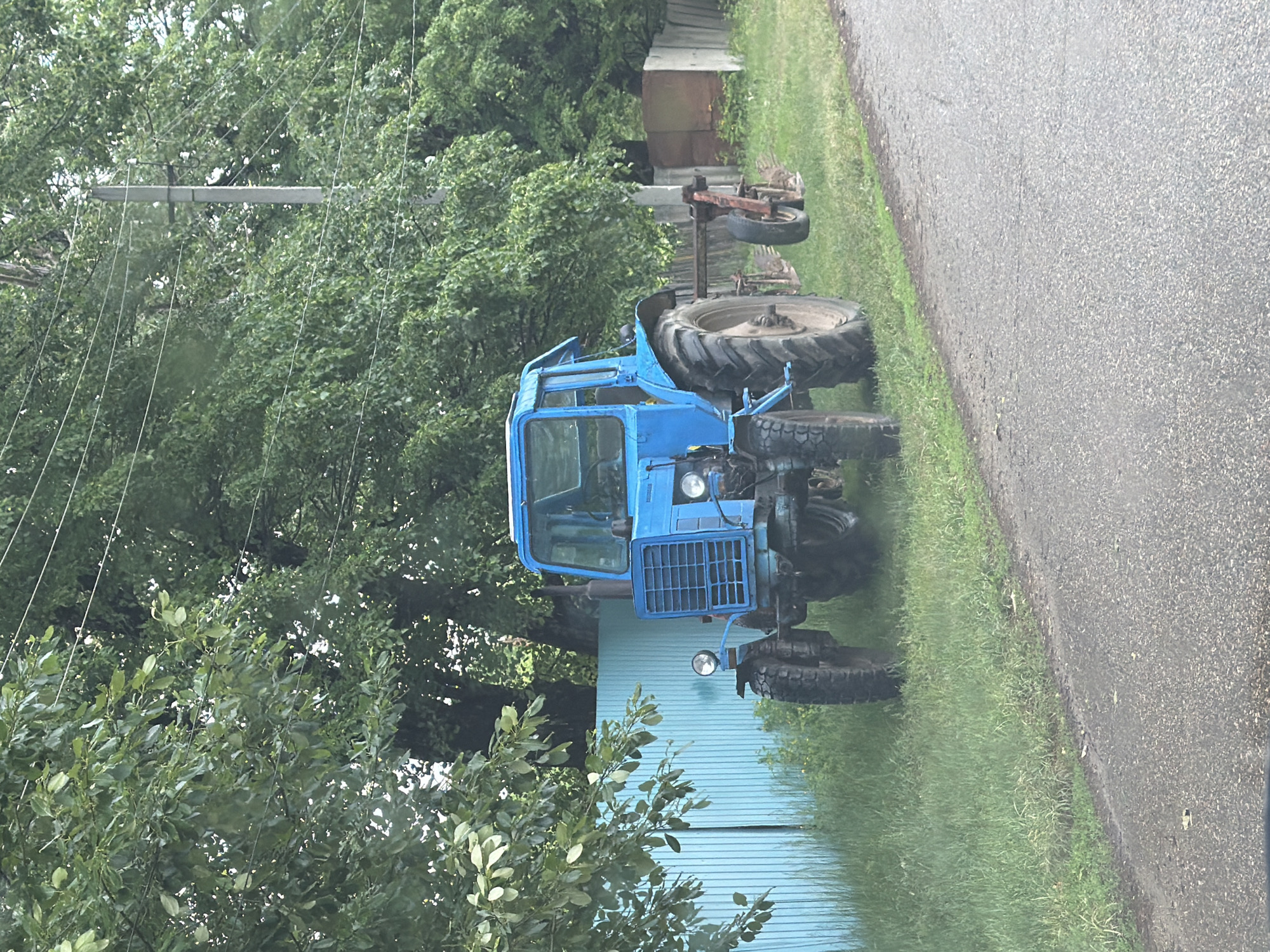 This forensic image captures a static, wide-angle view of a rural scene in Pronkino, Russia, under overcast or dappled light conditions. The primary subject is a faded blue agricultural tractor, likely a MTZ model, positioned on a grassy verge immediately adjacent to an asphalt road that occupies the lower portion of the frame. The tractor is equipped with a front grille, headlights, and a vertical exhaust pipe on the right side. A two-furrow plow, with one wheel lifted from the ground, is attached to the rear of the tractor. Behind the tractor, a light blue corrugated metal fence extends horizontally from the left, partially obscured by green foliage. Further right, a less distinct fence line constructed from concrete slabs or weathered wood is visible, with a dilapidated, reddish-brown structure or material behind it. A dense backdrop of vibrant green deciduous trees fills the upper half of the image, with a prominent utility pole, likely concrete, vertically bisecting the background on the right. Multiple thin power lines are stretched across the frame, connected to the utility pole and disappearing into the tree canopy. No individuals are present in the image, and no specific actions or interactions are depicted. The overall environment suggests a well-maintained but rural roadside setting.