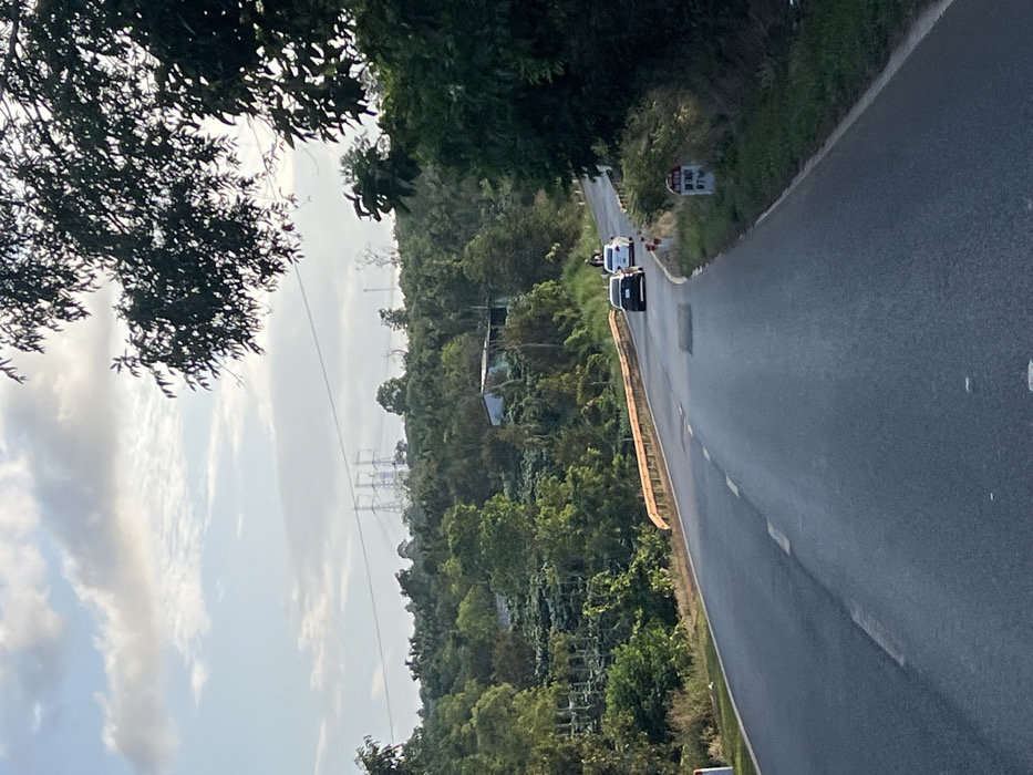 This image captures a view along an asphalt road in Ea Ksô, Vietnam, under a partly cloudy sky. The road, featuring visible lane markings and several patched areas, curves gently to the right as it extends into the distance. On the left side of the road, an orange-yellow metal guardrail outlines a bridge. Beyond the bridge, the landscape consists of densely forested hillsides, with some cultivated areas visible as rows of smaller trees or plants. A small, light-colored, single-story building is nestled among the trees on a hillside to the left of the road.

Three vehicles are present on the road, traveling away from the camera: a dark-colored sedan is closest to the foreground, followed by a person riding a white motorbike, and a white sedan further ahead. On the right side of the road, a white and red kilometer marker post is positioned on the grassy shoulder, displaying "ĐN BNT" and "07 KM". In the far background, multiple large, grey metal electricity transmission towers rise above the distant, tree-covered hills. The scene is illuminated by diffused daylight, indicative of morning or late afternoon.