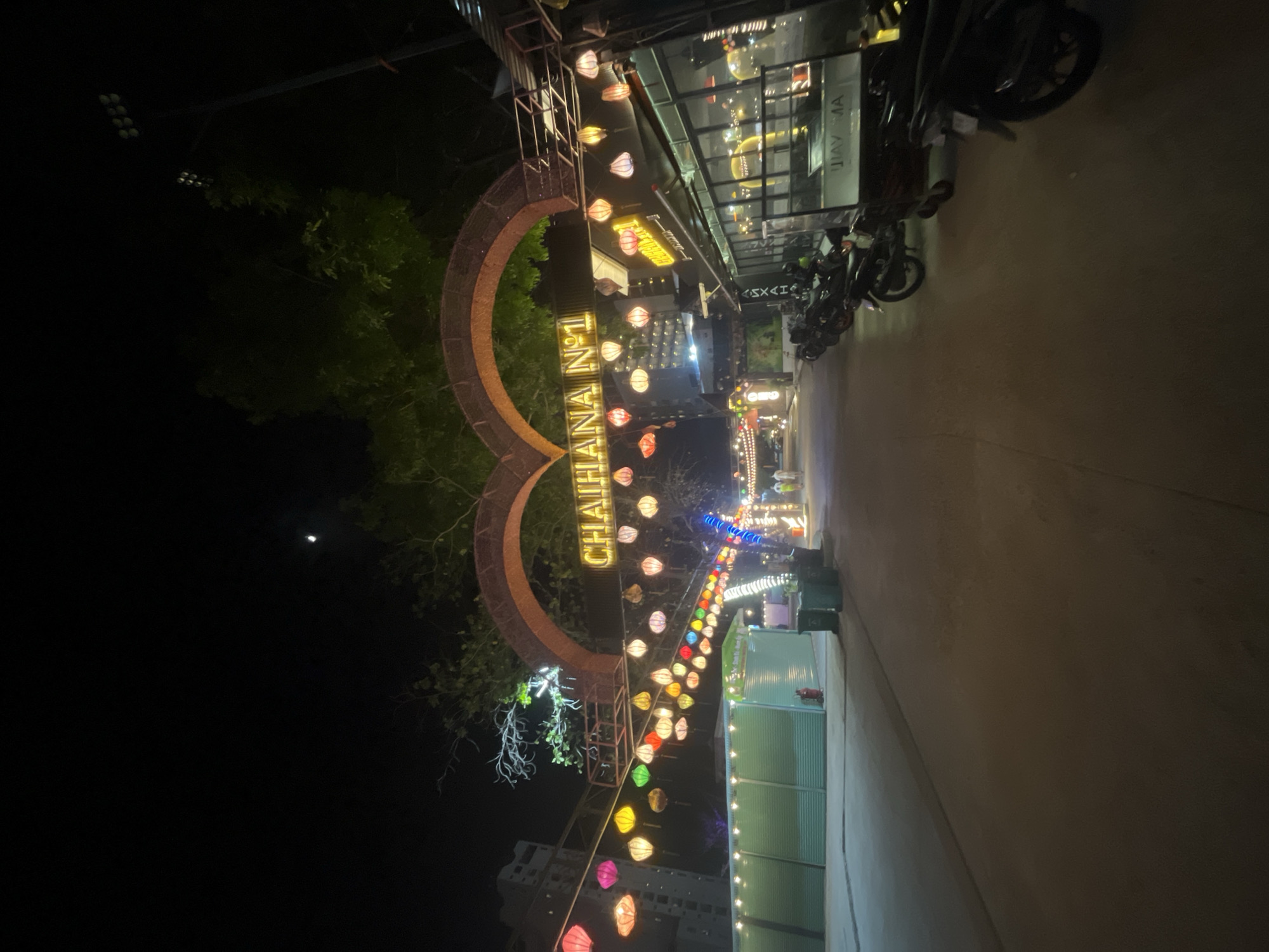 The image shows a nighttime street scene in Nha Trang, Vietnam, featuring an arched gateway illuminated by bright yellow neon lights spelling out "CHAIHANA N°1". Numerous colorful paper lanterns are strung across the street beneath the arch, creating a festive atmosphere. To the left, a building with white siding is visible, and further down the street, a row of green stalls or shops lines one side. On the right, a building with large glass windows appears to be a restaurant or bar, with several motorcycles parked in front. The street is paved and appears to be a pedestrian walkway or a quiet road. There are a few people visible in the distance, walking or standing. The overall impression is of a lively, well-lit entertainment or dining district at night.