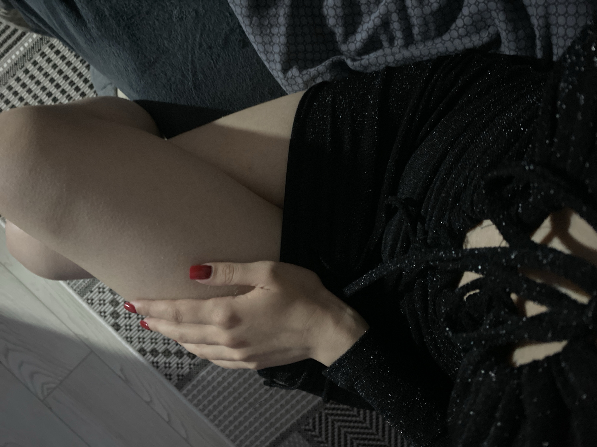 The image presents a close-up, high-angle view of a person, primarily showing their legs and one hand. The person is wearing a black, sparkly garment, likely a dress or top, which features intricate cut-out or laced details on the right side. Their left hand, adorned with bright red nail polish, is resting on their left leg. The skin tone is fair.
The setting appears to be indoors, characterized by dim lighting that suggests evening or low-light conditions. A soft glow illuminates the person's skin. In the background, a patterned rug or carpet with a black and white geometric design is visible on the lower left. Above this, a dark grey, textured fabric, possibly a blanket or upholstery, is present, partially covered by another grey fabric with a subtle circular pattern. A light-colored wooden floor is visible along the bottom and left edge of the frame. The overall composition suggests a relaxed or posed posture within a domestic environment. There is no visible text in the image.