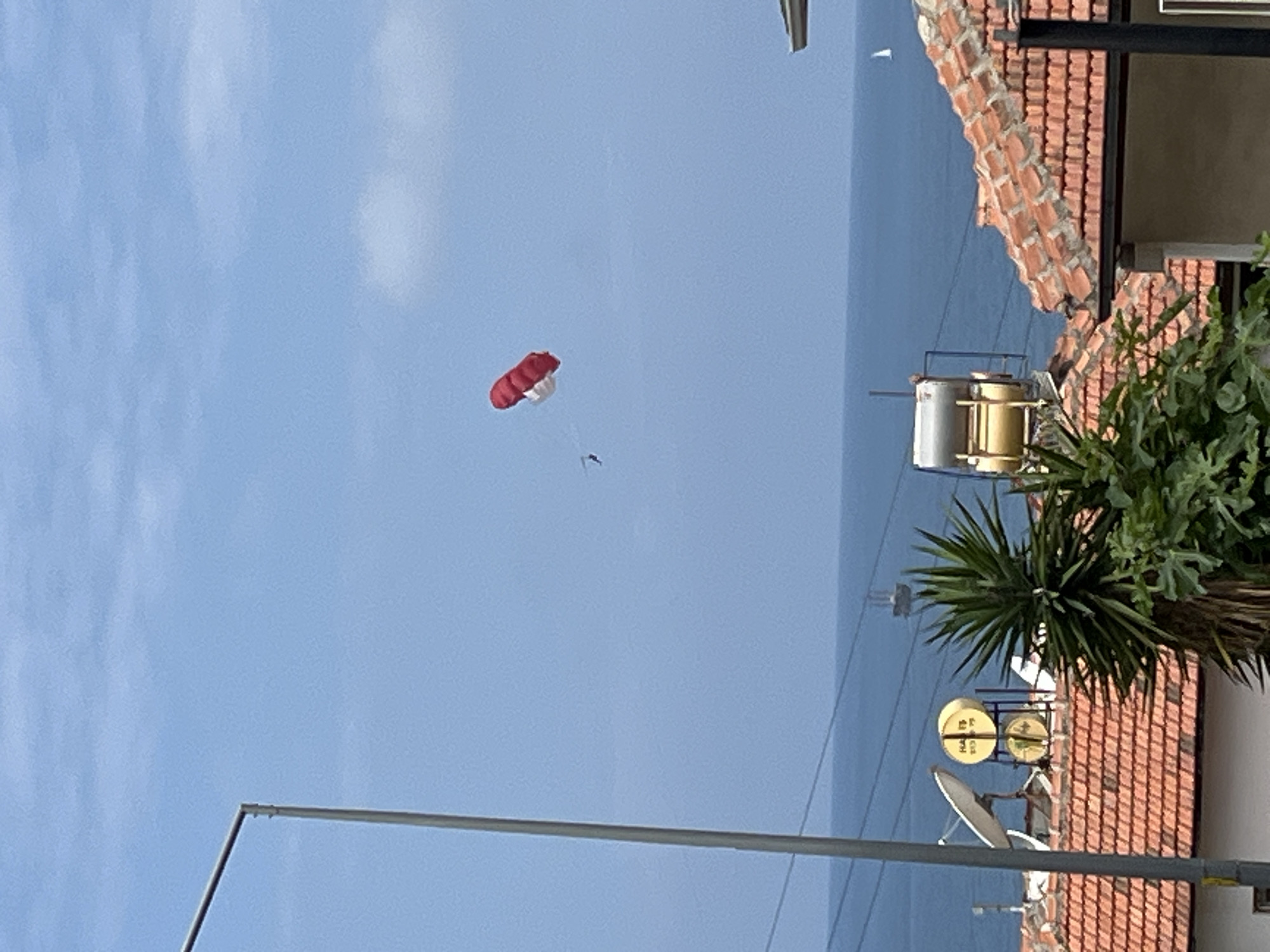 A parachute, red and white in color, is visible in the sky. Below it, a person hangs in the air. In the background, a building with a red tile roof and green plants nearby is apparent. A satellite dish is attached to the building. A street light is visible near the bottom of the image. The photo was likely taken in Alanya, Turkey.