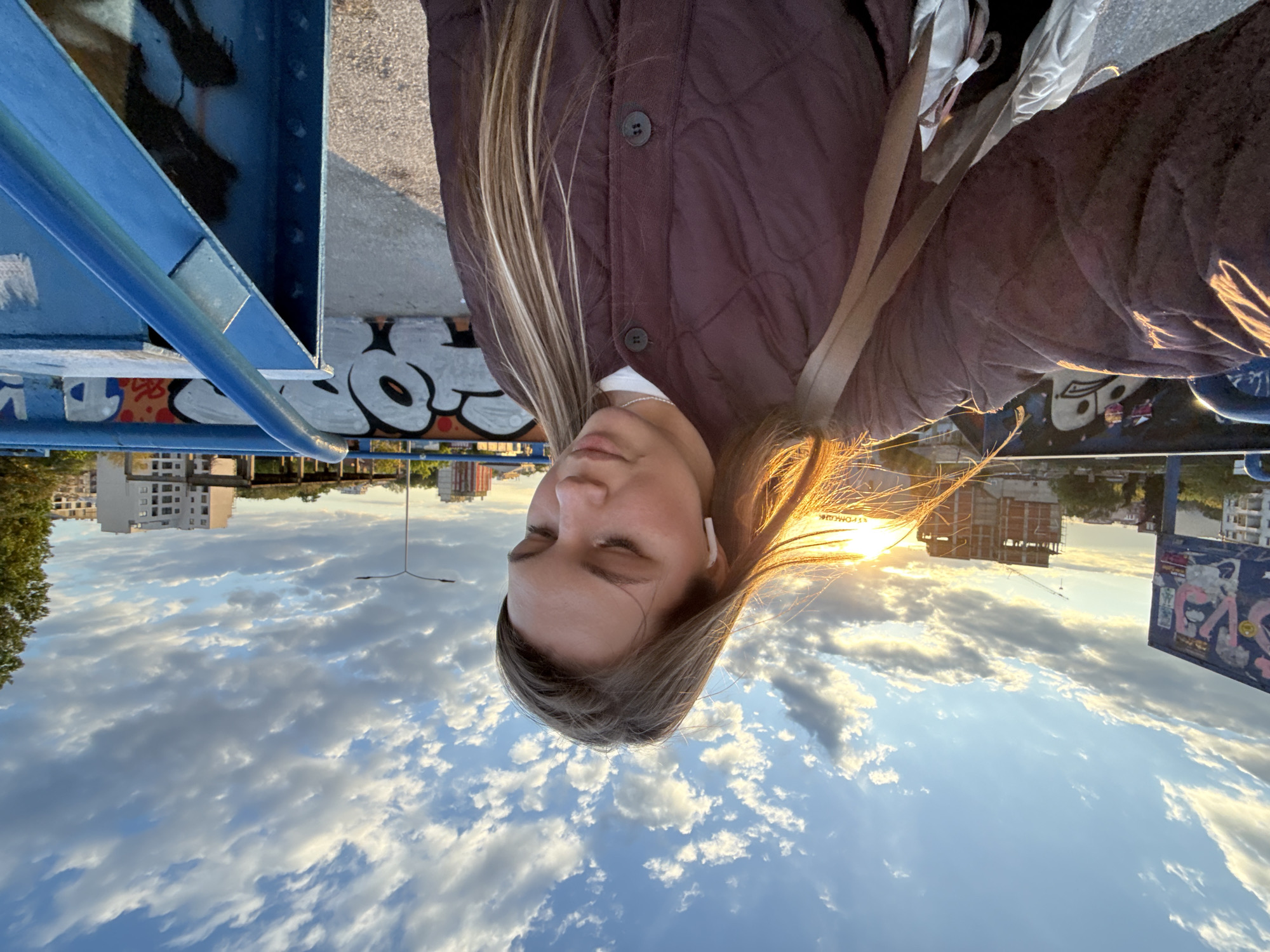 The image captures a serene outdoor scene, featuring a young woman in the foreground against an urban backdrop during the golden hour.
The primary subject is a young woman, likely in her late teens or early twenties, with long, highlighted light brown hair that glows warmly from the sunlight. She is wearing a dark maroon quilted jacket over a white top and has white wireless earbuds in her ears. Her eyes are closed, and her head is tilted slightly downwards, suggesting a moment of peace or reflection. A silver chain is visible around her neck.
She appears to be standing on or near an elevated structure, possibly a bridge or overpass, indicated by a prominent blue metal beam and rail on the left side of the frame. Underneath this structure, large white bubble-letter graffiti with black outlines is visible. The background reveals an urban landscape with several buildings, including a white multi-story apartment complex and another building under construction. A lamppost stands in the mid-ground.
The time of day is either sunrise or sunset, characterized by the low sun on the right, which bathes the scene in a warm, golden light and casts a glow on the woman's hair. The sky is partly cloudy, with scattered cumulus clouds adding texture to the blue expanse. On a distant blue sign or board, faint Cyrillic text can be discerned, possibly "ЕФРОМЕЧИК", which aligns with the given location context of Vozdovac Urban Municipality, Serbia. The overall mood is tranquil and contemplative.