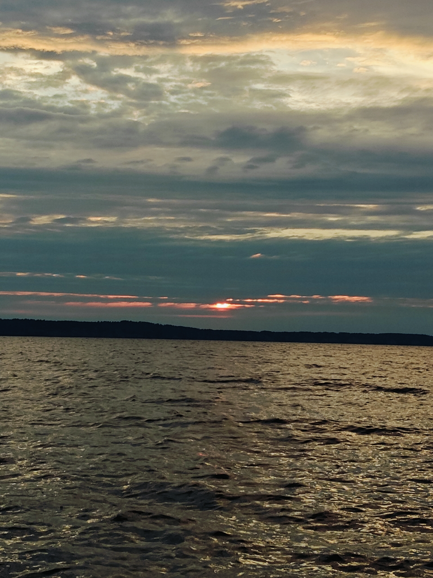The image shows a body of water, likely a river or lake, at sunset. The sky is filled with clouds, some of which are tinged with pink and orange from the setting sun. A dark, treeline is visible on the horizon, separating the water from the sky. The water itself is choppy, with reflections of the sky and the setting sun visible on its surface. The scene is serene and tranquil, suggesting a peaceful end to the day. Based on the provided context of Chaykovsky, Russia, this image is likely depicting the Kama River or Votkinsk Reservoir, both significant bodies of water in the vicinity of the city.