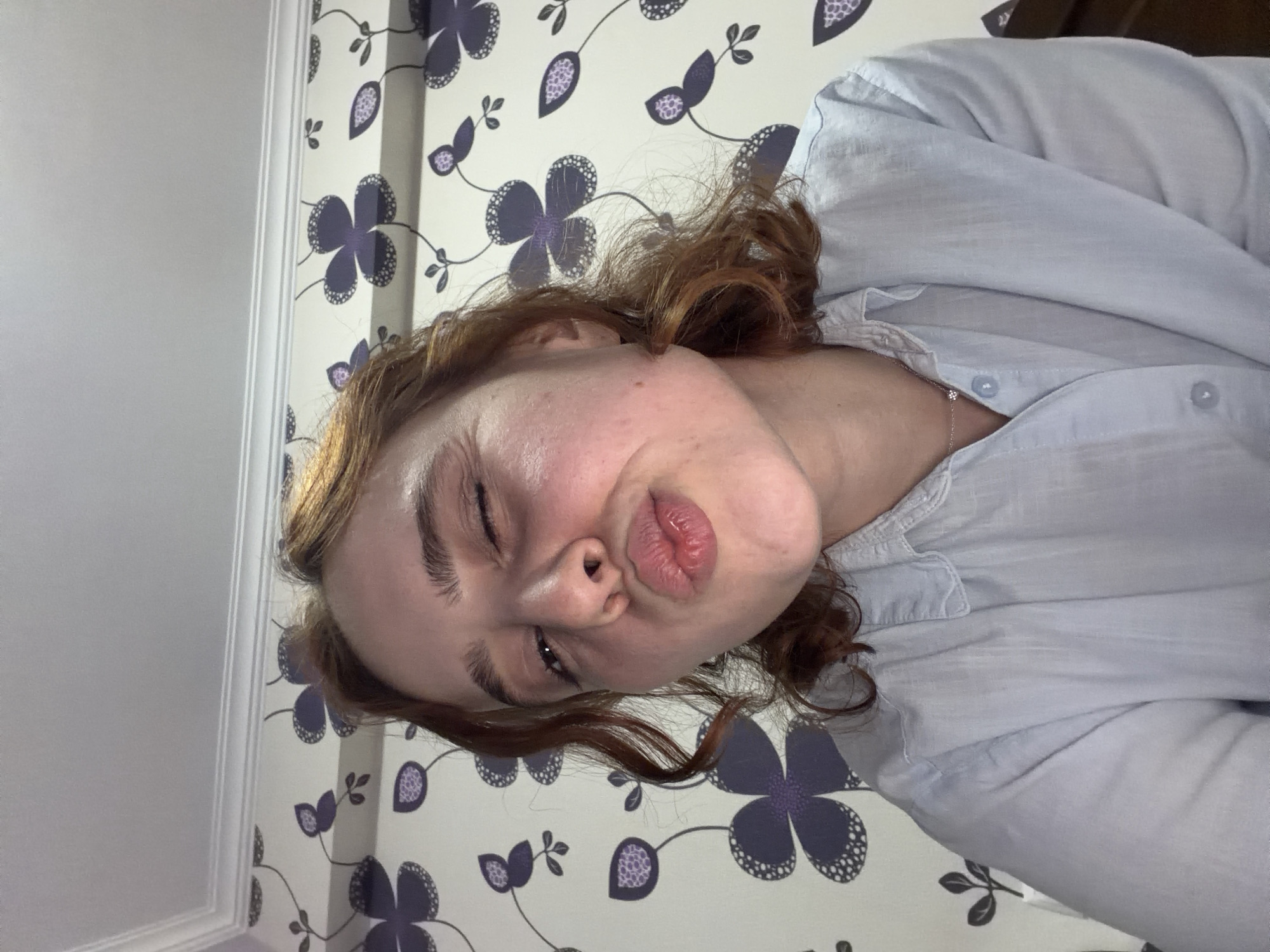 A pale-skinned person with reddish-brown curly hair poses for a close-up selfie in what appears to be an indoor setting. The person is wearing a light blue button-up shirt and possibly a necklace. The background consists of wallpaper with a repeating floral pattern featuring purple flowers with black polka dots. Part of a white wall or trim is visible on the left side. Given the information available, it is impossible to ascertain the location in the city of Tbilisi, Georgia.