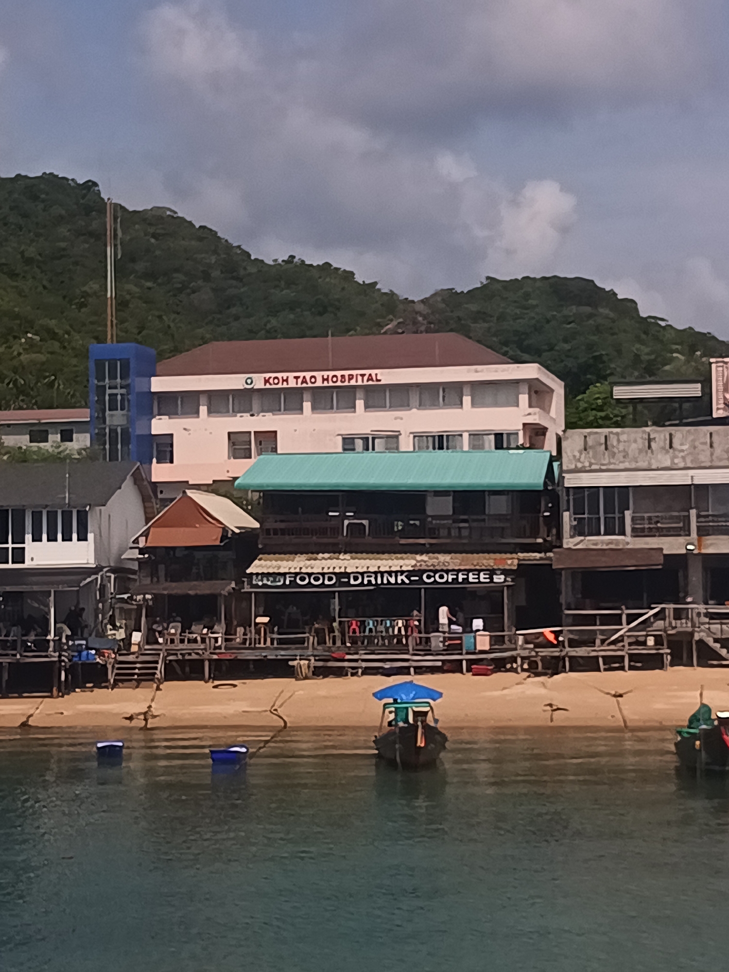 The image depicts a waterfront scene in Tambon Ko Tao, Thailand. A hospital building is visible in the background, with a sign reading "KOH TAO HOSPITAL." A series of wooden structures, likely businesses offering food, drink, and coffee, line the beach. The beach itself is sandy, with several small boats floating in the water near the shore. There are wooden docks and platforms extending from the buildings. People appear to be present in the businesses, though they are somewhat indistinct. The environment includes a hillside covered with trees and a cloudy sky.