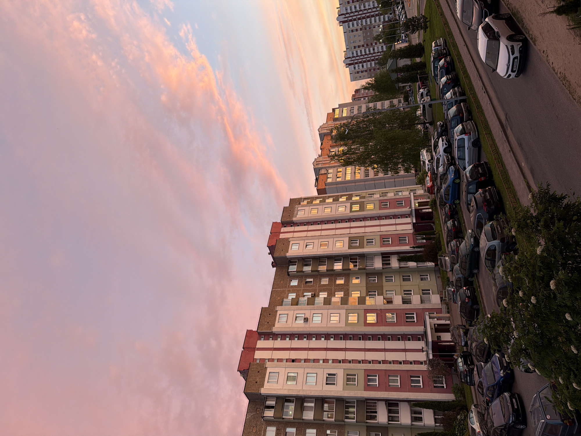 The image shows a cityscape at sunset. On the right side of the image, there's a road lined with parked cars. To the left of the road are apartment buildings of varying heights and colors. The sky is filled with pink and orange clouds, suggesting a sunset. There are no people visible in the image.