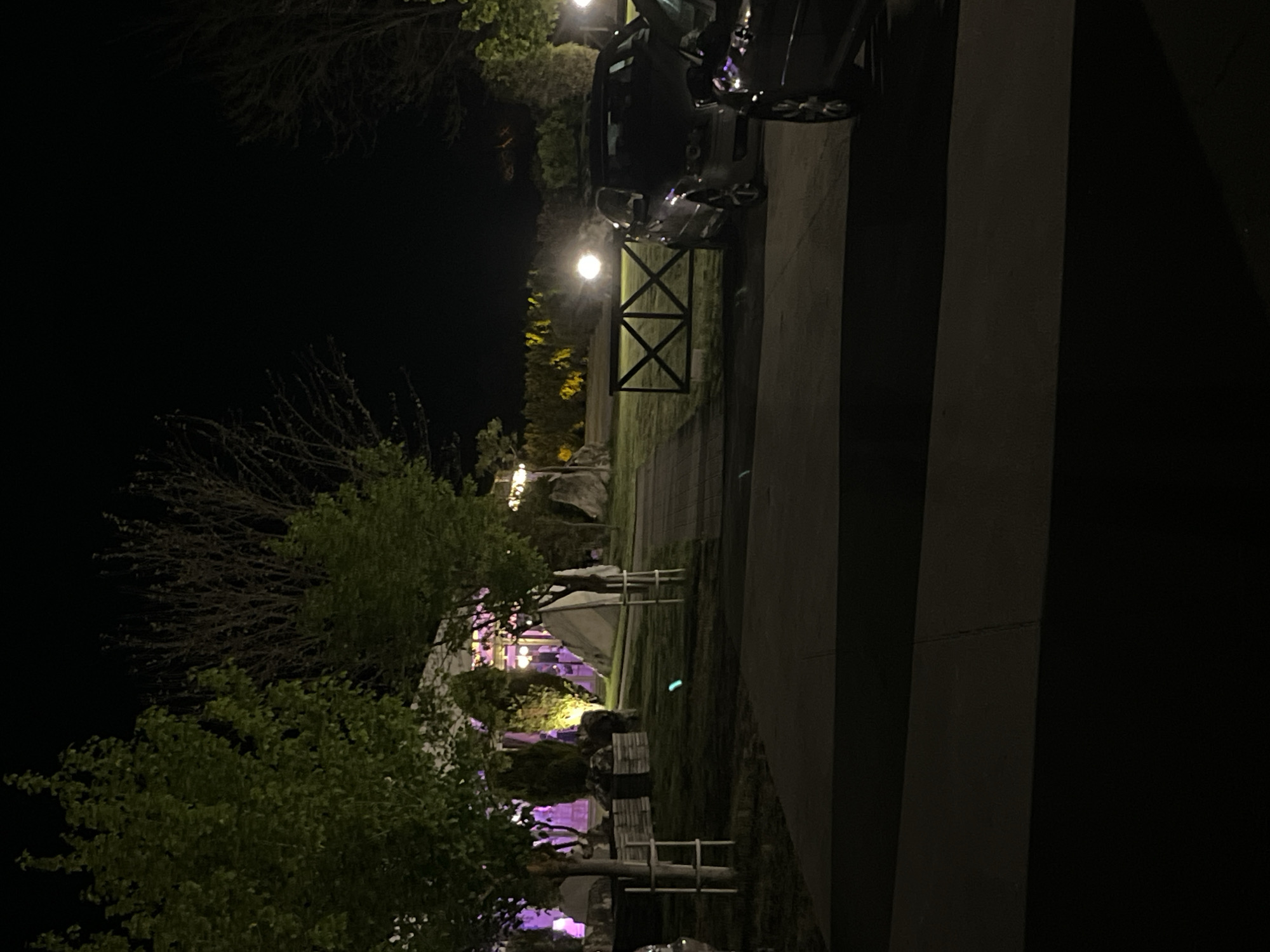 At night, a dark SUV sits next to a walkway with vertical concrete features and an open lawn. Beyond the SUV are lit, manicured trees. In the background, illuminated white tents and purple lights can be seen in Koprivlen, Bulgaria.