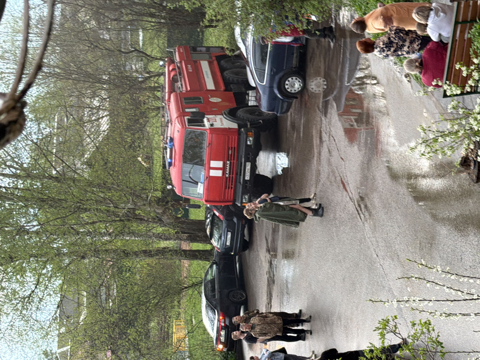 The image shows a street in Reshetnikovo, Russia, on a wet day. A red fire engine, a KAMAZ, is parked on the road alongside several cars. People are standing around the vehicles, some observing, while others are sitting on a bench nearby. Trees line the street, their leaves just emerging in spring. The road surface is wet and reflective.