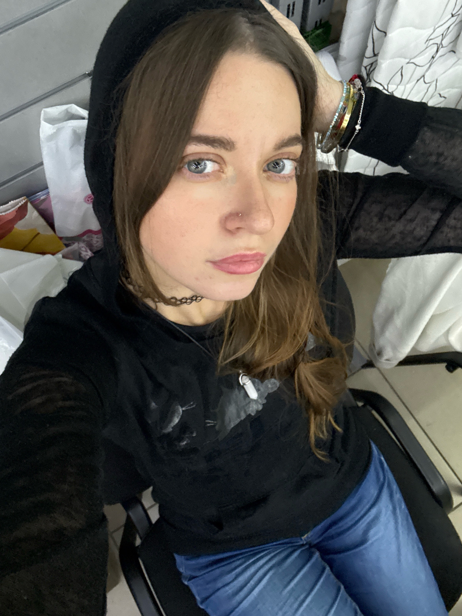 A young woman with long brown hair and blue eyes is taking a selfie. She is wearing a black hooded sweatshirt and blue jeans. She has on a black choker necklace and several bracelets on her left wrist. She is sitting in a black chair in what appears to be a home environment. The background includes bags and white fabric. The location is described as being in Chaikovsky, Russia, but this cannot be visually confirmed from the image alone.