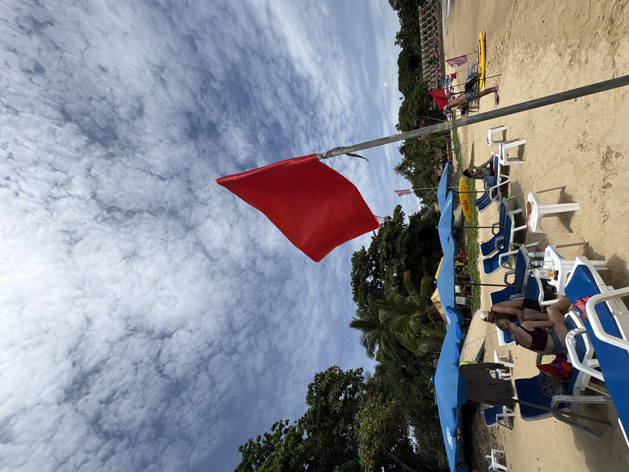The image depicts a beach scene on a cloudy day at Karon Beach, Thailand. A prominent red flag, indicating dangerous sea conditions, flutters from a pole in the foreground. The sandy beach is lined with blue lounge chairs and umbrellas. Several people are visible on the beach, some relaxing on chairs, while others appear to be engaged in activities near the water. A yellow kayak and a "RESCUE" sign are also present, suggesting lifeguard presence. Lush green trees and palm trees form a backdrop to the beach, with buildings visible in the distance.