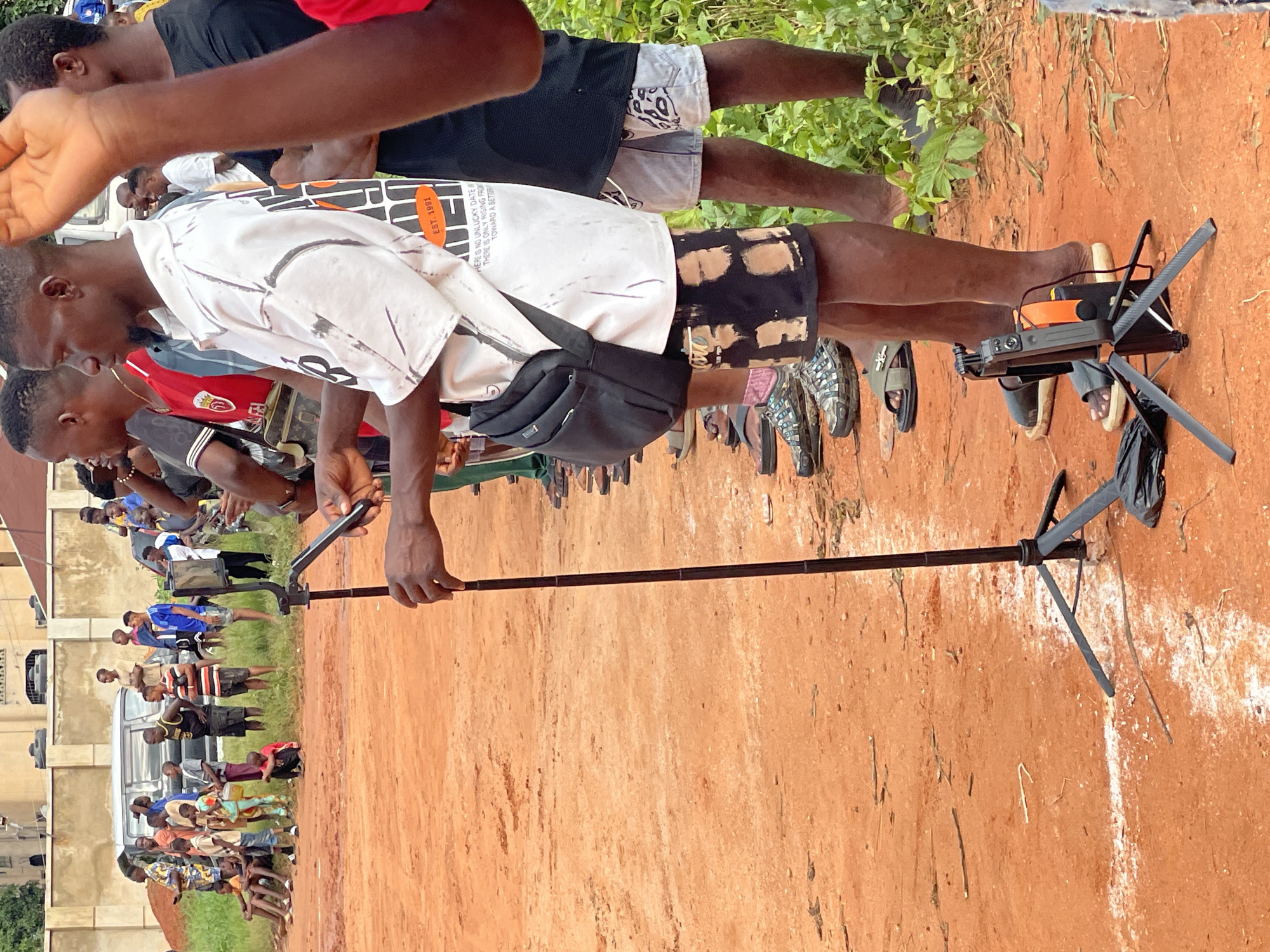 In an outdoor, dusty, orange-colored environment with some patchy green vegetation on the right, a group of people, predominantly young males, are gathered. In the foreground, a tripod with a mounted device, possibly a camera or phone, stands on the ground. A person in a white, short-sleeved shirt with a black graphic design and a black crossbody bag is holding a dark-colored object, likely a smartphone, and looking down at it. To their right, another person in a black t-shirt and light-colored patterned shorts stands. A thin, dark-colored stick or pole is positioned vertically in the scene, held by a hand visible in the upper right of the frame. In the background, a beige building with dark window frames is visible, along with a silver van and a crowd of onlookers, including some seated individuals. The overall scene suggests an event or gathering.