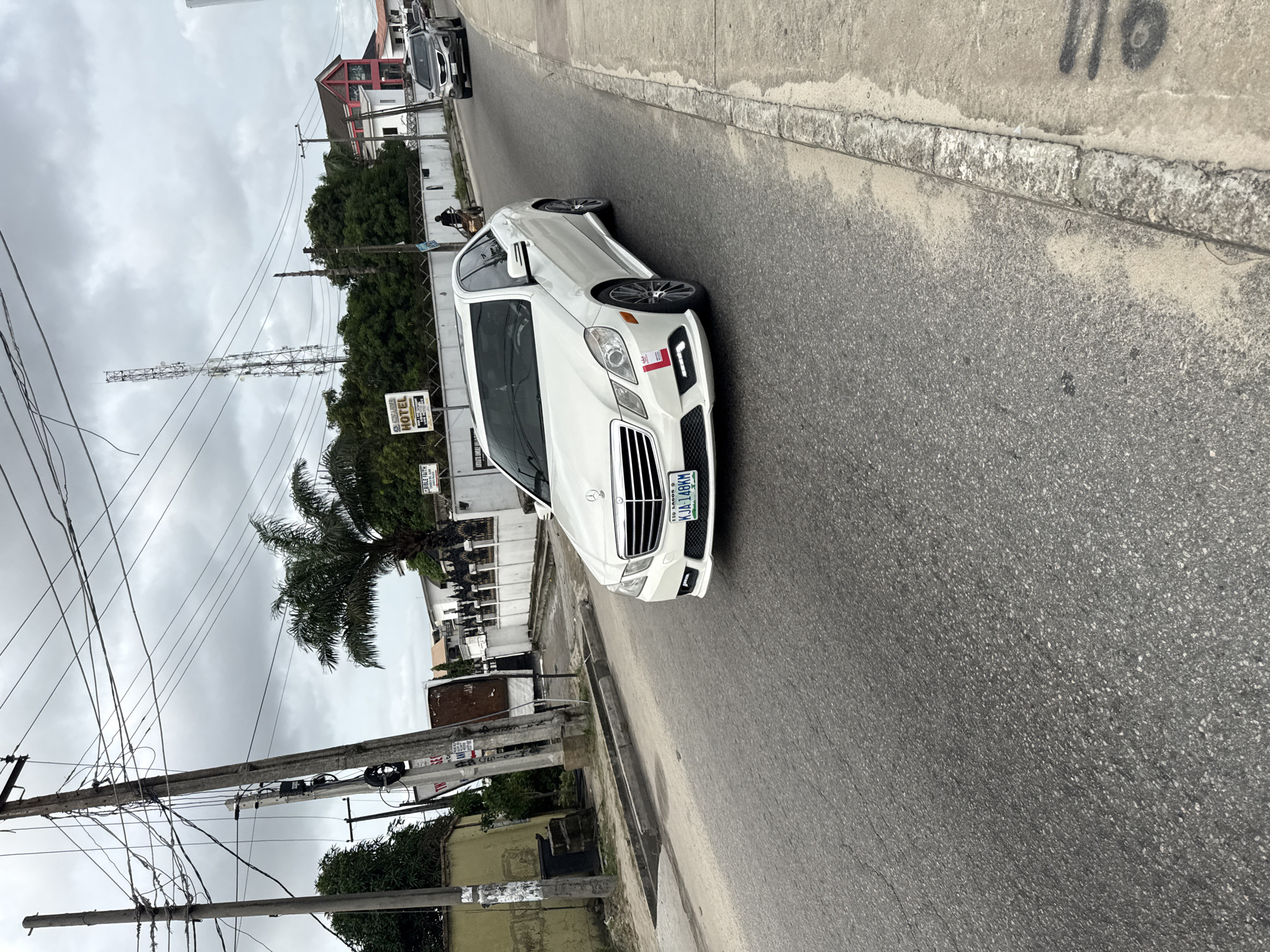 A white Mercedes-Benz E-Class sedan is parked on the side of a paved road in Ado, Nigeria. The car has a Nigerian license plate reading "KJA 148 KM". In the background, a communication tower rises above trees and buildings, including a sign indicating a hotel. Electrical wires crisscross the sky above the street. A motorcycle with a rider is parked further down the road. The sky is overcast.