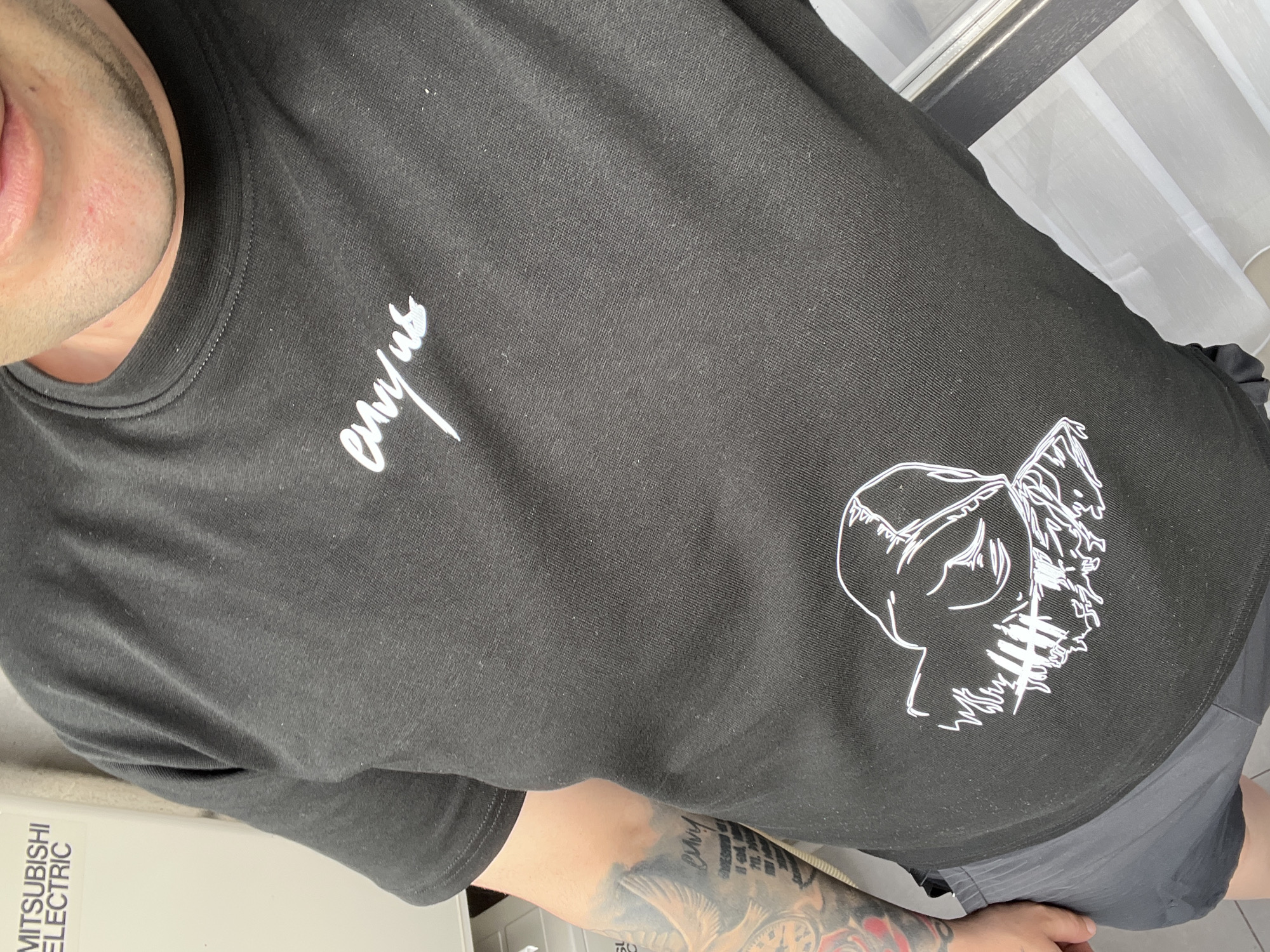 A person is wearing a black t-shirt with white designs, including cursive writing near the shoulder and an outline of a human head near the bottom. The person also has a tattoo on their arm. The setting appears to be indoors, near a window with white curtains, and adjacent to a white Mitsubishi Electric unit. The specific location within Varna, Bulgaria, cannot be determined from this image.