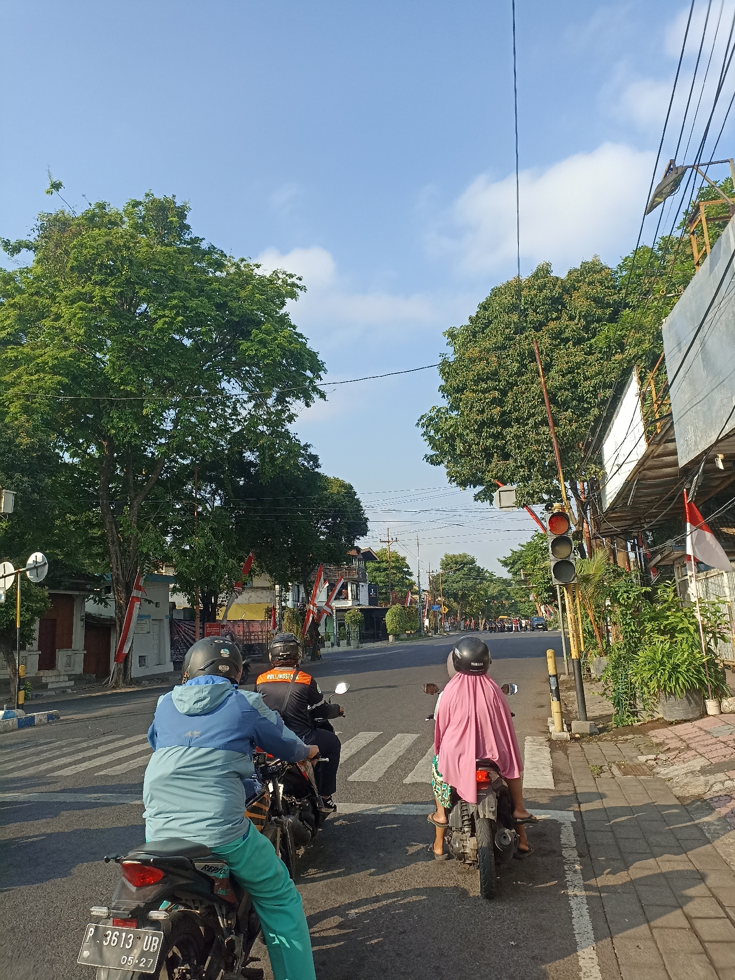 The image depicts a street scene in Mojopanggung, Indonesia, under a clear blue sky with scattered clouds. In the foreground, two motorcycles are stopped at a crosswalk. The first motorcycle has two occupants. The rider, facing away from the camera, wears a blue and green jacket and green pants, with a black helmet. The passenger, also facing away, is covered in a pink headscarf and wears patterned shorts. The second motorcycle, further ahead, has a single rider wearing a black and orange jacket with the word "POLISI" visible, and a black helmet.
The road is paved, with clear white crosswalk markings. To the right of the road is a sidewalk with textured paving stones. A traffic light is visible, displaying a red light. Lush green trees line the street, with some buildings and shops visible in the background. Indonesian flags are displayed on some of the buildings. The overall impression is a typical urban streetscape in Indonesia during daylight hours.