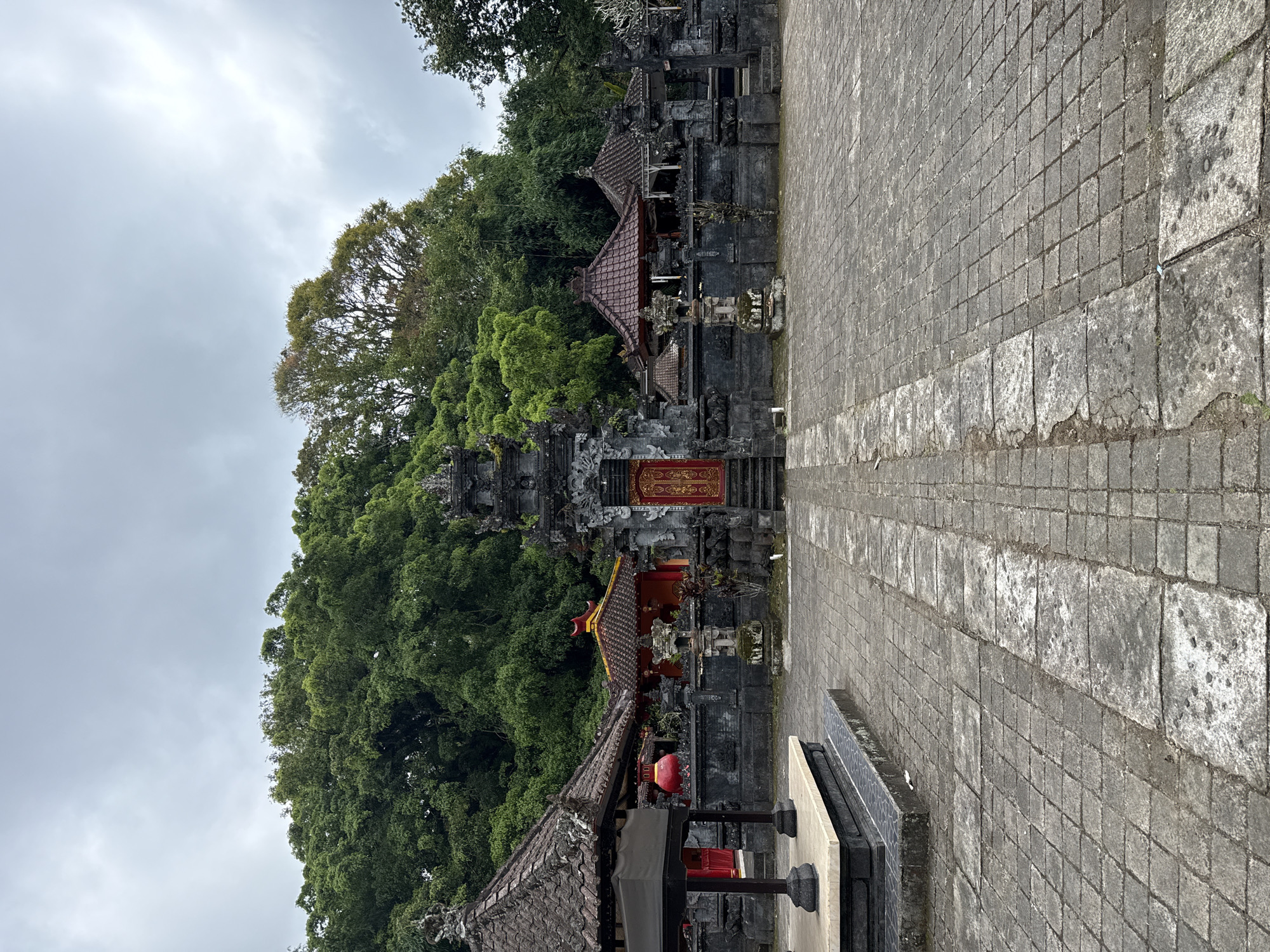 The image captures a serene view of a traditional Balinese temple complex in Baturiti, Indonesia, under an overcast sky. To the left, a lush, tree-covered hillside forms a dense green backdrop, with various shades of foliage. To the right, a wide, grey stone-paved pathway or courtyard extends into the mid-ground, featuring square and rectangular stone blocks that show signs of age and wear.
Between the vibrant greenery and the paved area stand several ornate temple buildings constructed from dark stone, featuring distinctive multi-tiered roofs covered with reddish-brown terracotta tiles. A prominent feature is a central gate, possibly a candi bentar, adorned with striking red panels and intricate gold ornamentation. This gate displays decorative, illegible script or calligraphy in gold.
Further into the complex, other temple structures with similar architectural styles and tiled roofs are visible amidst the trees. One structure in the foreground, adjacent to the pathway, appears to have red, lantern-like decorations hanging beneath its roof. The overcast sky suggests soft, diffused daylight, lending a tranquil atmosphere to the cultural site. No people are visible, emphasizing the stillness and spiritual essence of the location.