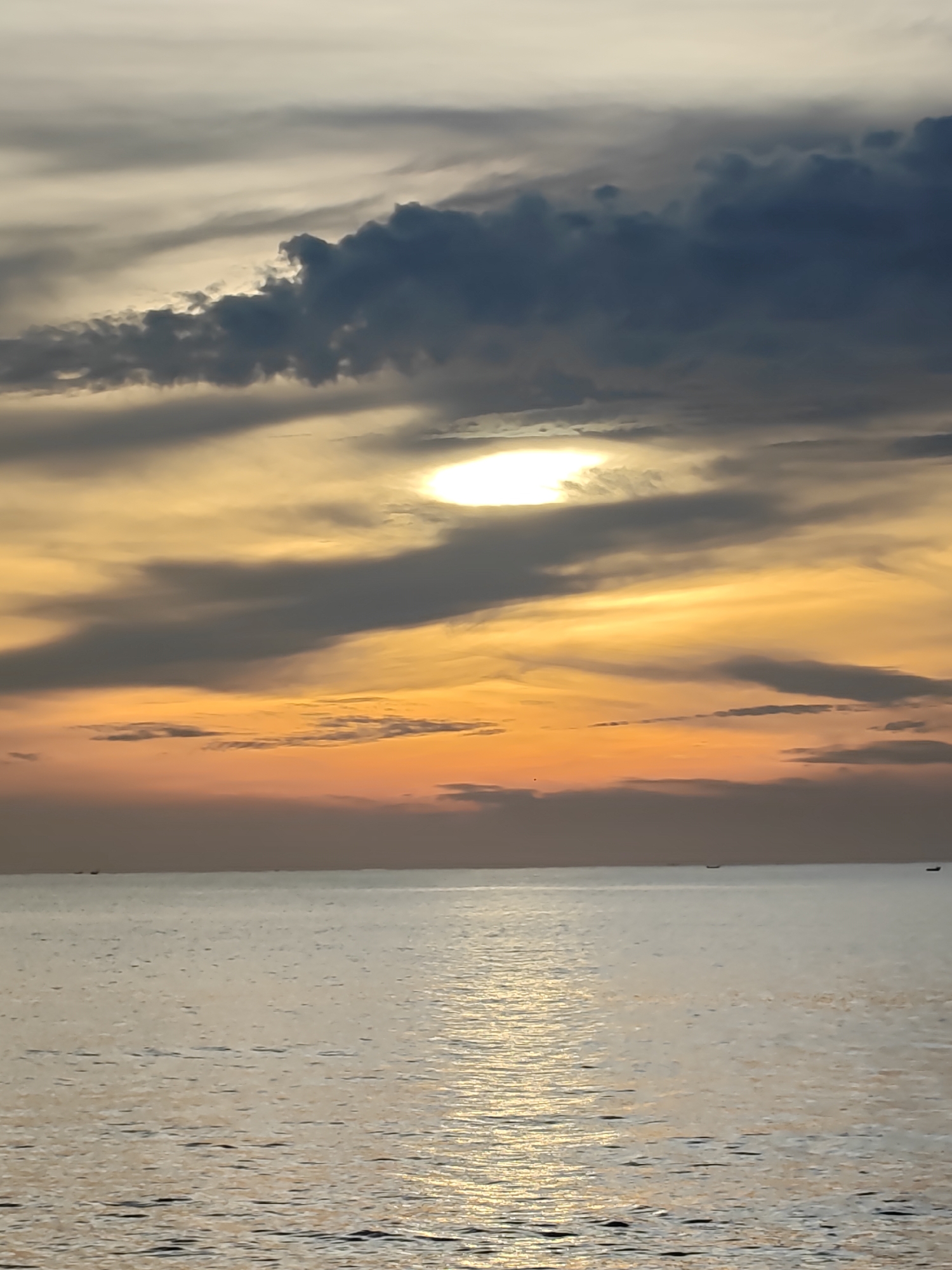 This vertical image depicts a coastal seascape in Lhokseumawe, Indonesia, captured during either sunrise or sunset. The sky occupies the upper two-thirds of the frame, characterized by a complex cloud formation. In the upper-mid section, a bright, circular luminary (presumed to be the sun) emits intense white light, partially obscured by a prominent band of dark, dense stratocumulus clouds. Surrounding and beneath these darker clouds, the sky exhibits a gradient of colors, transitioning from light grey and white at the very top to vibrant golden yellows and fiery oranges closer to the horizon. Streaks and wisps of altocumulus clouds are visible throughout the colored portions of the sky.
The lower third of the image presents a calm ocean surface, which reflects the overhead light, forming an elongated path of shimmering golden-white light extending from the foreground towards the horizon. The water itself appears predominantly muted grey-silver with subtle ripples. Several small, dark, indistinct silhouettes, consistent with distant fishing boats or vessels, are visible on or just above the flat horizon line, particularly towards the left and right edges of the frame. No human figures or discernible landmasses are visible.