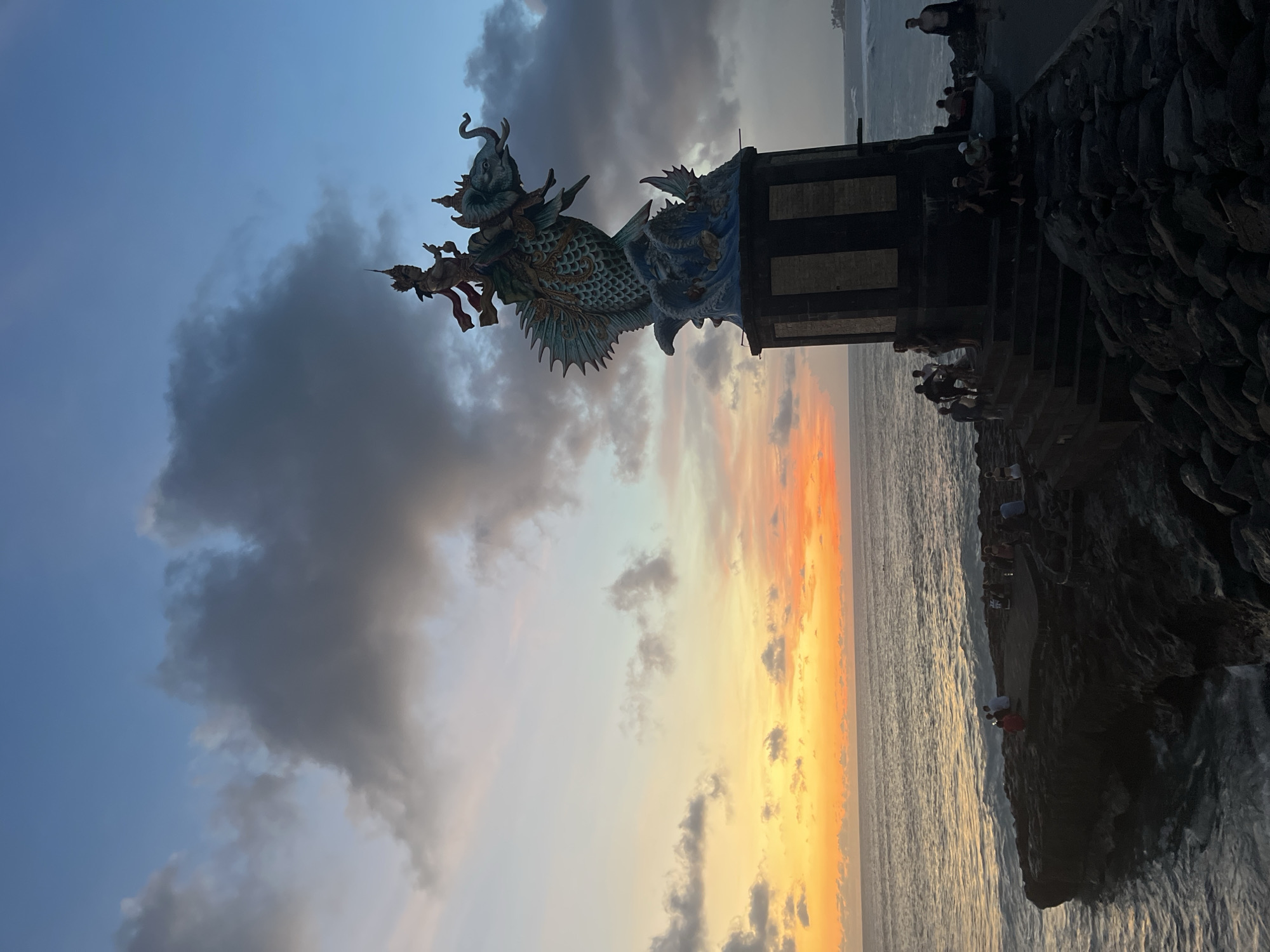 The image captures a dramatic coastal scene at sunset in Canggu, Indonesia. A large, ornate dragon statue stands on a stone pillar atop a rocky outcrop overlooking the ocean. The statue features intricate details, including scales and multiple figures. The sun is setting over the ocean, casting an orange and yellow glow across the water and sky. Dark clouds contrast with the vibrant sunset colors. Several people are gathered on the rocky outcrop and stairs surrounding the statue, appearing to be tourists or locals enjoying the view. The ocean is visible, stretching towards the horizon. The photo is taken from a slightly low angle, emphasizing the height of the statue and the vastness of the sky.