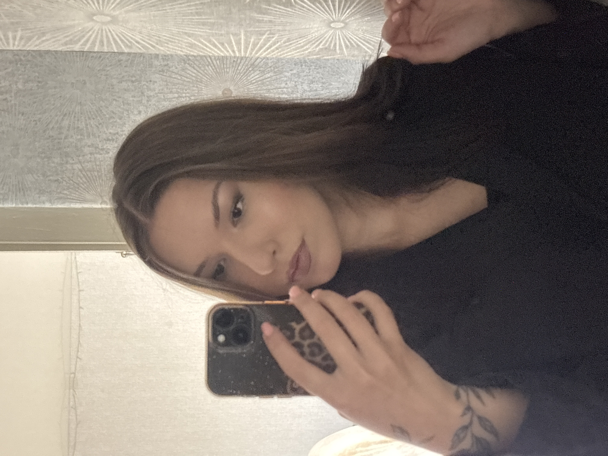 A woman is taking a selfie in a dimly lit room. She is holding a smartphone with a leopard print case in her right hand, capturing her reflection in a mirror. The woman has long, dark brown hair and is wearing a black garment. Her left arm, visible in the lower left of the frame, has a tattoo of leaves and vines. The background appears to be a bathroom or dressing area, with a textured wallpaper featuring a sunburst pattern on the right side and a white, semi-transparent curtain or shower screen on the left. The lighting is soft, and the overall impression is of a private, indoor setting. The location is identified as Inskoy, Russia.