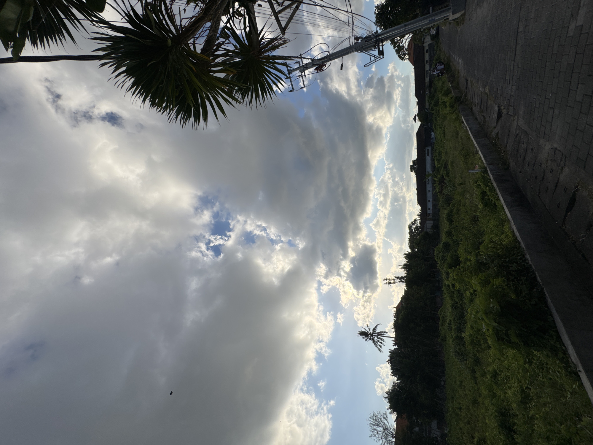 The image displays an outdoor scene in Kuta, Indonesia, under a dynamic, partly cloudy sky. The upper half is dominated by large, dark grey cumulus clouds interspersed with patches of bright white clouds and clear blue sky, suggesting late afternoon light with the sun obscured but illuminating cloud edges. In the upper left quadrant, a small, dark, rectangular object, possibly a drone or kite, is visible against the clouds. The upper right features the silhouetted fronds of a palm tree and a grey utility pole laden with multiple power lines, extending diagonally downwards.
The mid-ground presents a dense line of tropical trees and shrubs, mostly dark green, forming a natural barrier. Behind this foliage, parts of several low-rise buildings with traditional Indonesian-style roofs are discernible. A prominent, solitary palm tree stands silhouetted against a bright patch of sky towards the middle left.
The foreground is divided into two distinct areas. On the left, a large, unkempt vacant lot is covered in dense, overgrown green vegetation. A low, grey concrete wall separates this lot from a paved road on the right. This road is constructed from dark grey interlocking bricks and shows signs of wear. The road appears to gently ascend into the distance. In the far distance on the paved road, two individuals are observed, one appearing to be on a scooter or motorcycle, with another person nearby.
