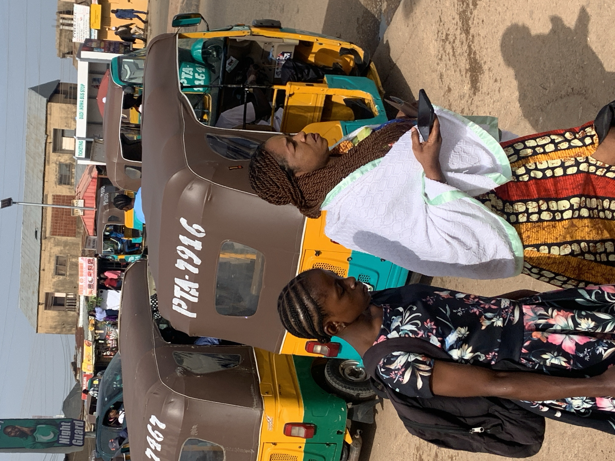 Two women and a younger girl are standing in a dusty street in Gura Top, Nigeria, with several auto-rickshaws visible. The younger girl, on the left, has her hair styled in thin braids and is wearing a dark floral print dress and a black backpack. She is looking down and to her left. To her right, an adult woman is wearing a white shawl with light green trim over a colorful patterned skirt. She is holding a dark-colored mobile phone in her right hand and looking to her right. Behind them, several auto-rickshaws, commonly known as Keke Napeps, are parked. One has "PTA-1916" written on its side, and another has "PTA 1159" visible. The background shows buildings, some with corrugated metal roofs, and a billboard with "Night Guard" text and a man's image. The ground is a mixture of dirt and small stones. The sky is clear and blue.