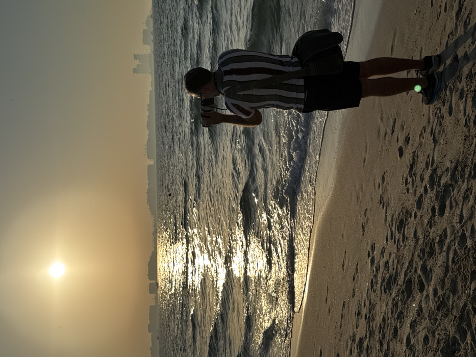 A male individual, viewed from behind, stands on a sandy beach in Dubai, United Arab Emirates. He is actively photographing the seascape with a black DSLR camera fitted with a long telephoto lens, held to his eyes. He wears a vertically striped shirt, dark shorts, and dark athletic shoes; a dark shoulder bag is slung across his body. The foreground features wet and dry sand marked with footprints. Gentle waves lap the shore, creating a foamy edge. The sea extends across the midground, intensely reflecting the bright, circular sun positioned in the upper left of the hazy, golden sky. A small, dark buoy floats in the water towards the left midground. The distant horizon reveals a low, hazy silhouette of numerous urban structures and buildings, characteristic of Dubai's coastal development or artificial islands. The overall illumination is consistent with golden hour.
