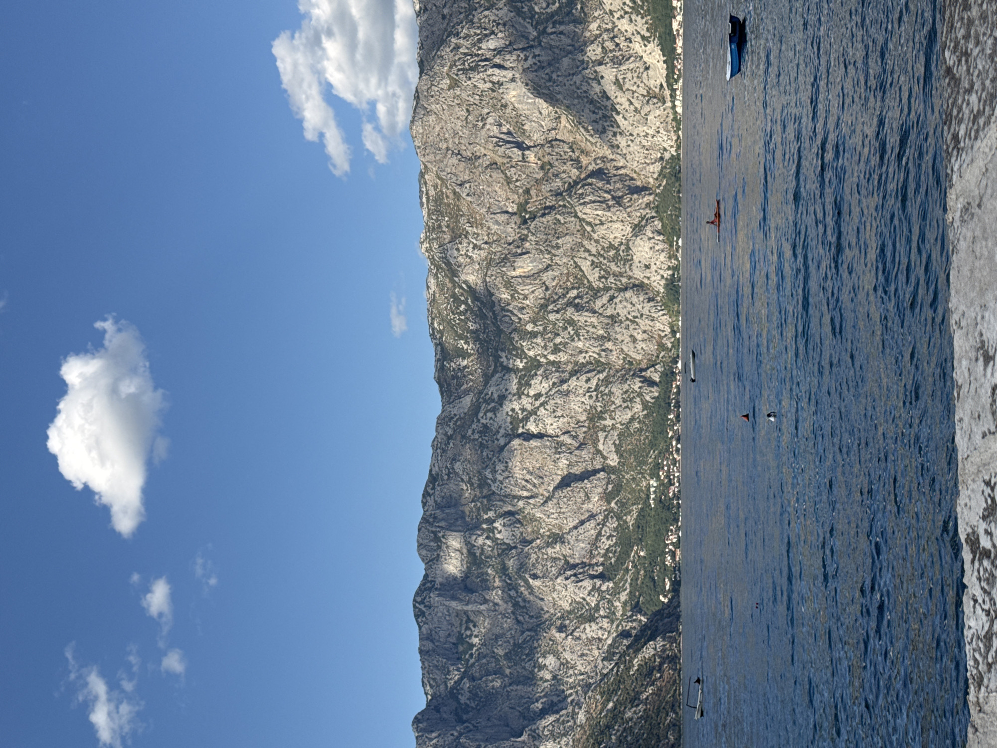 The image captures a serene scene in Prčanj, Montenegro, showcasing the dramatic mountainous backdrop of the Bay of Kotor. The vast, deep blue water occupies the majority of the frame, with gentle ripples indicative of a calm day. In the foreground, a textured, light-colored concrete structure, possibly a quay or walkway, is visible, hinting at the edge of the land.
Several small boats are scattered across the water. To the far left, a white boat is moored, its hull and the silhouette of a person within clearly discernible. Further into the bay, a small, dark-hulled boat is visible. Slightly to the right and closer to the viewer, a red kayak with a person seated in it is navigating the water. To the right, a small blue boat is at anchor. Two buoys, one red and one orange, are also present in the water, marking the area.
The middle ground is dominated by a densely vegetated coastline, with scattered buildings nestled amongst the greenery, suggesting a settlement along the bay. The background features a monumental, rugged mountain range that rises steeply from the coastline. The mountains are characterized by their grey, rocky surfaces, marked by deep fissures and crevices, with sparse patches of vegetation clinging to the slopes. The peaks are sharply defined against a clear, bright blue sky, which is adorned with a few fluffy white clouds. The overall impression is one of natural grandeur and tranquility.