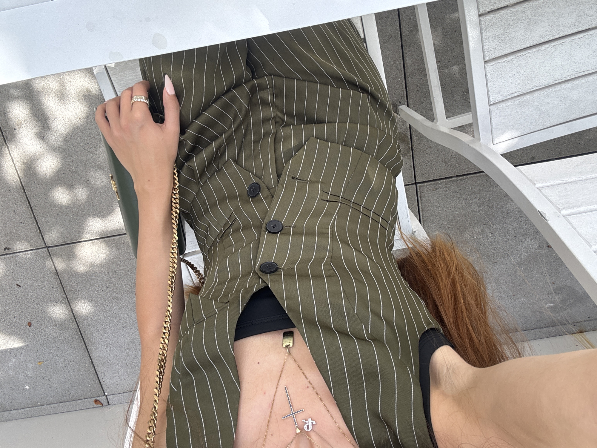 A woman's body, from the mid-chest down, is visible, angled to the right. She wears a green pinstripe blazer over a black garment. A gold chain necklace with a cross and a small charm hangs around her neck. Her right hand rests on her lap, adorned with two rings. Her left arm is bent, and her hand is on her stomach. Her long, auburn hair is visible on the lower right side of the image. The woman is seated on a white chair next to a white table, outdoors, on a tiled patio. The sun casts shadows from an overhead structure.