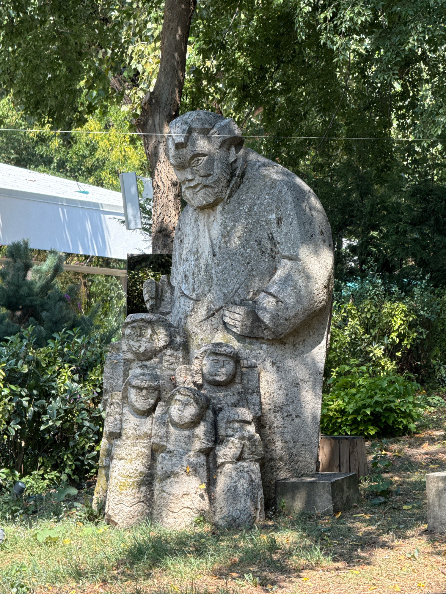 This is a full shot of a stone sculpture depicting a large man standing over a group of smaller figures. The sculpture is located in Bucharest, Romania, in the Carol I Park. The large figure has a stern expression and is wearing a long robe. The smaller figures appear to be children, with their faces carved in a simplified style. The sculpture is set in a natural environment with trees and foliage surrounding it. The lighting suggests it is daytime. There are no visible actions or interactions occurring in the scene.