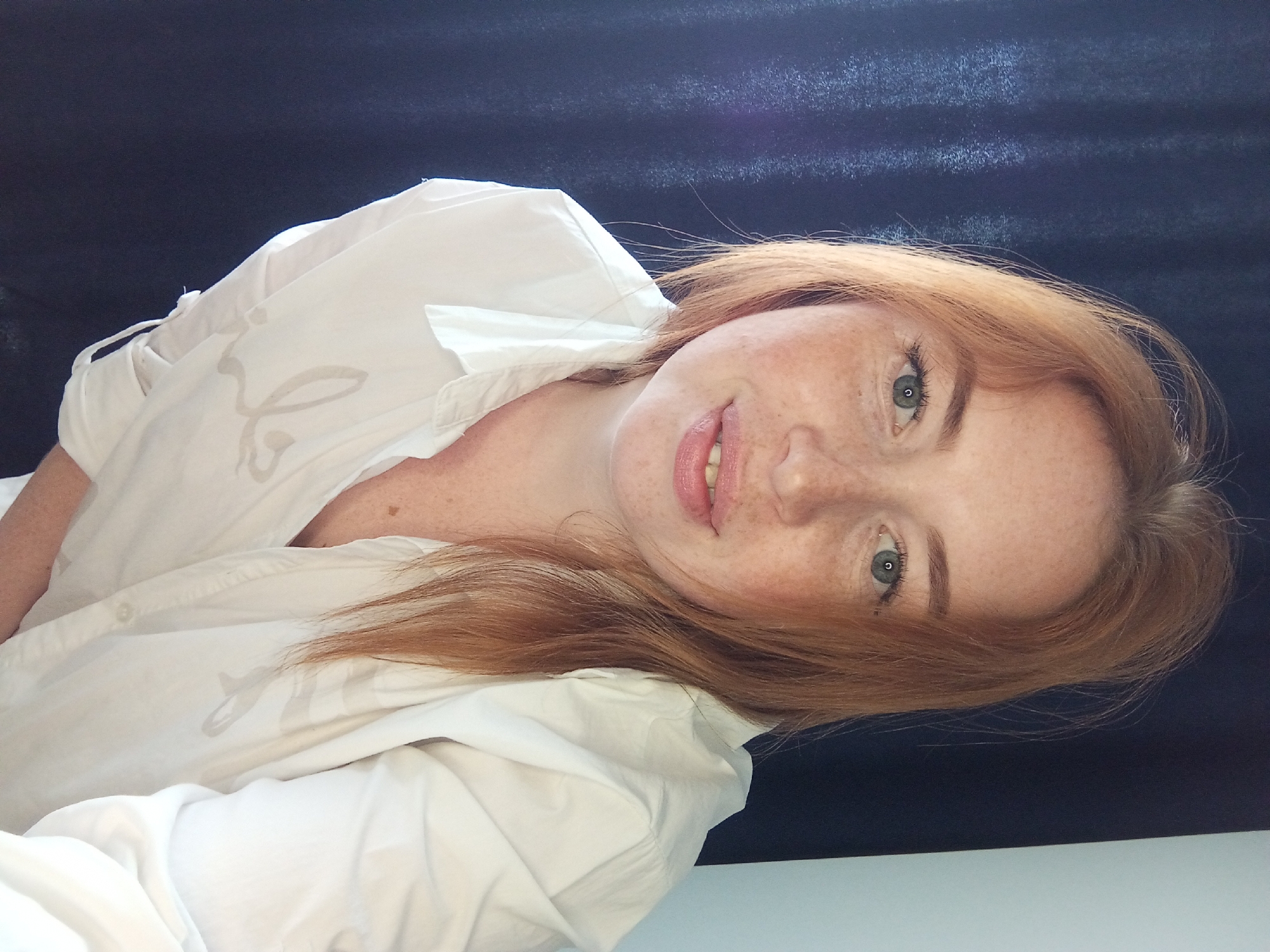 A light-skinned woman with freckles and reddish-blonde hair is pictured from a close-up, slightly angled perspective. She is wearing a white button-down shirt, partially unbuttoned. The background appears to be a dark blue or black surface. The image does not contain information to support a location.