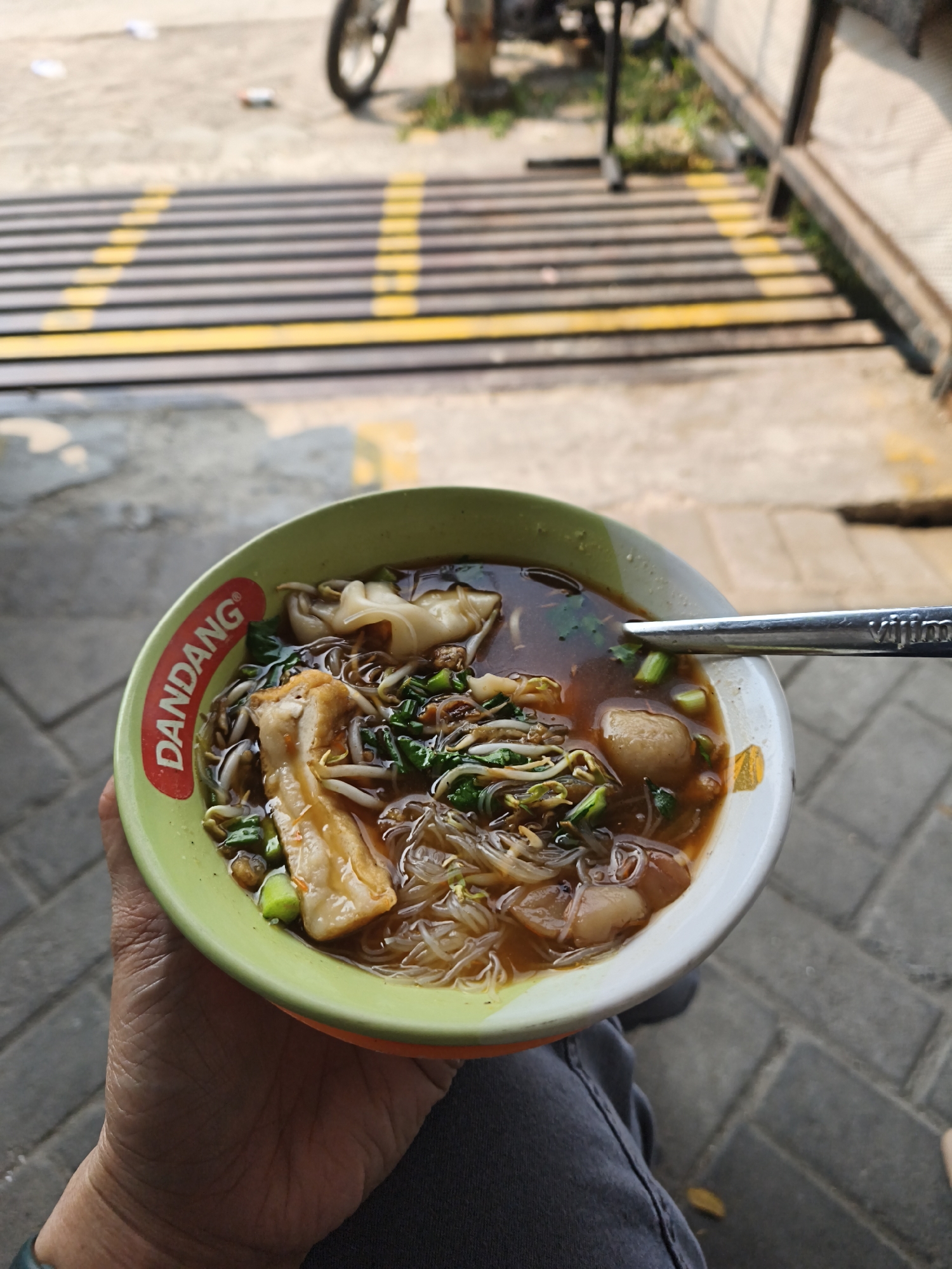 A close-up, high-angle shot shows a bowl of Indonesian noodle soup being held by a person's hand. The bowl, green on the outside with a white interior, has the brand "DANDANG" printed in red on its rim. The soup contains noodles, bean sprouts, green onions, a piece of fried tofu, meatballs, and wontons in a rich broth. A silver spoon rests on the edge of the bowl. The background is out of focus, showing a paved surface with yellow and black striped markings and some vegetation, suggesting an outdoor setting, possibly a street or market in Bandar Lampung, Indonesia.