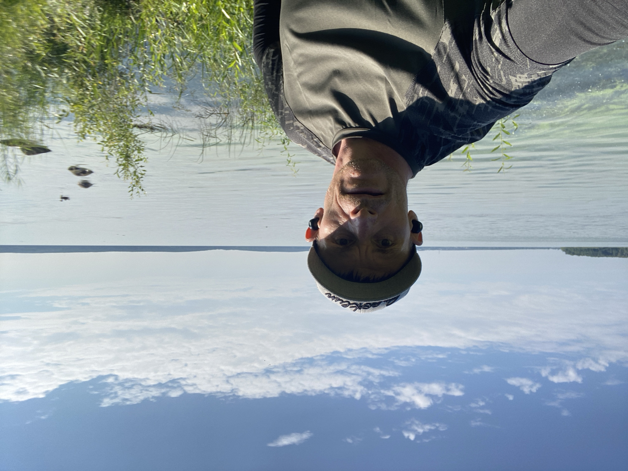 A man wearing a baseball cap and a long-sleeved olive green shirt is standing in front of a large body of water, likely a lake or reservoir, under a partly cloudy blue sky. He is looking at the camera and wearing earbuds. The water appears calm with some light green algae or vegetation visible near the shore. Tall green grass and some small bushes are growing along the water's edge in the foreground. A few ducks or birds are visible in the distance on the water, along with some rocks or small islands. The background shows a distant tree line and a faint horizon. The image appears to be a selfie taken outdoors during the daytime. The location is identified as the Yekaterinburg city, Russia, likely a natural area within or near the city.