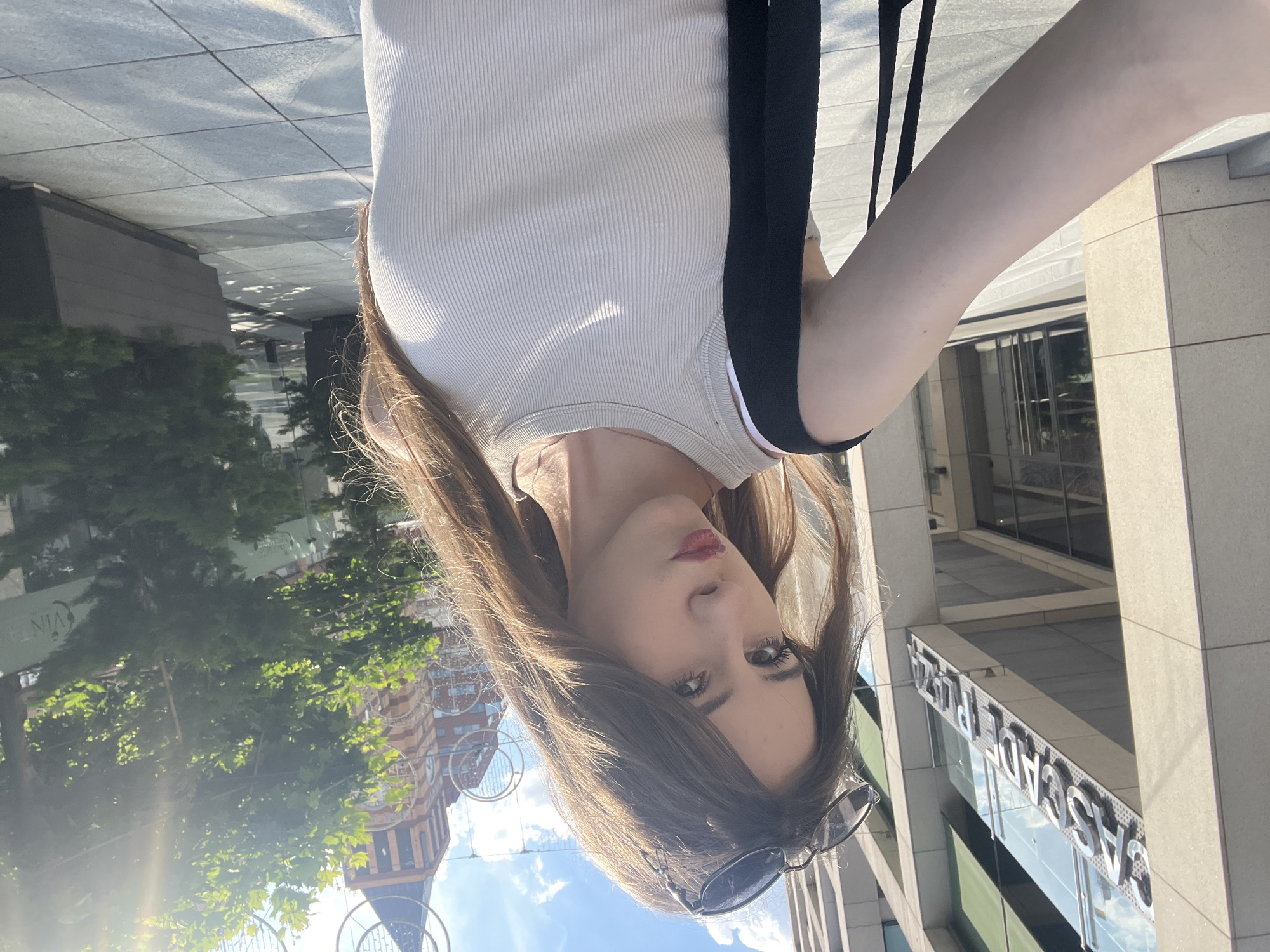 This media file shows a close-up, upside-down, portrait-oriented selfie of a young woman. She has long, brown hair, light-colored eyes, and is wearing a light-colored ribbed tank top with black straps. She is also wearing sunglasses pushed up on her head. The background suggests an urban outdoor setting.
The scene appears to be a sunny day, indicated by the bright light and shadows. In the background, a building with a modern facade and a sign that reads "CASCADE PLAZA" is visible. There are also trees with green foliage and a building with a distinctive red and brown brick facade, possibly a clock tower, which is a landmark in Dnipro, Ukraine. The ground appears to be paved with large, light-colored tiles, reflecting the sunlight.
The woman's expression is neutral, and the overall mood of the image is casual and informal, typical of a selfie taken outdoors. The time of day appears to be daytime, given the strong sunlight and shadows. There are no specific activities or events taking place other than the taking of the photograph.