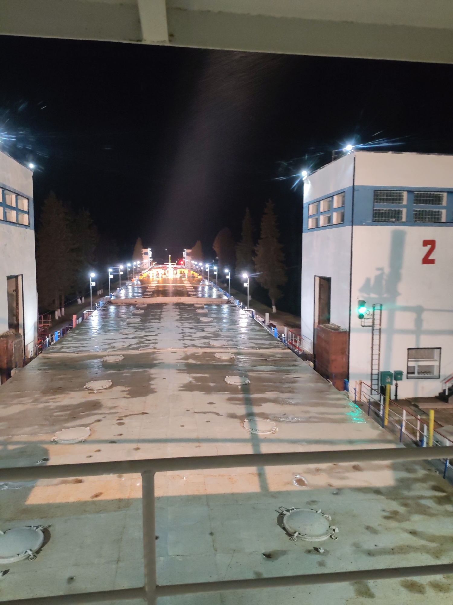 The image depicts a nighttime view of a canal lock in Белоусово, Russia. The scene is characterized by a concrete surface with several circular manhole covers in the foreground. The central area is illuminated by a series of evenly spaced streetlights lining the canal. On either side, there are buildings with blue and white paint schemes and visible windows. On the right-hand side, a large red number "2" is painted on the side of the building. A ladder and a traffic light indicating a green signal can also be seen near the building on the right. In the distance, lights suggest the presence of additional structures or features further along the canal. Trees are visible in the background. The overall impression is one of industrial infrastructure at night.