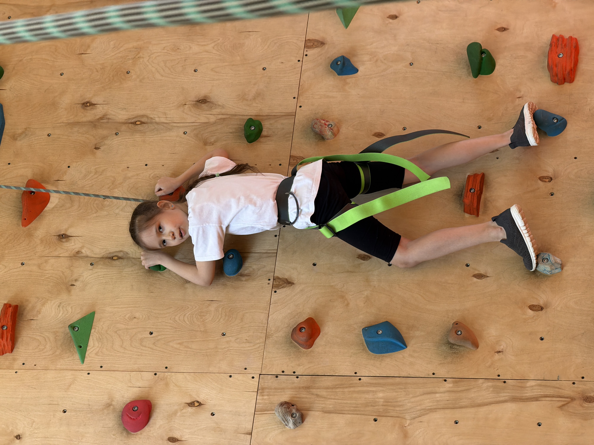 A young girl is positioned horizontally on a wooden climbing wall, wearing a white T-shirt, black shorts, and gray shoes. She is secured with a green and gray safety harness. Her feet are placed on climbing holds. The wall features an assortment of colored climbing holds in various shapes and sizes, attached with bolts. The background is the wooden surface of the climbing wall, marked with several bolt holes. The location is Izhevsk, Russia.