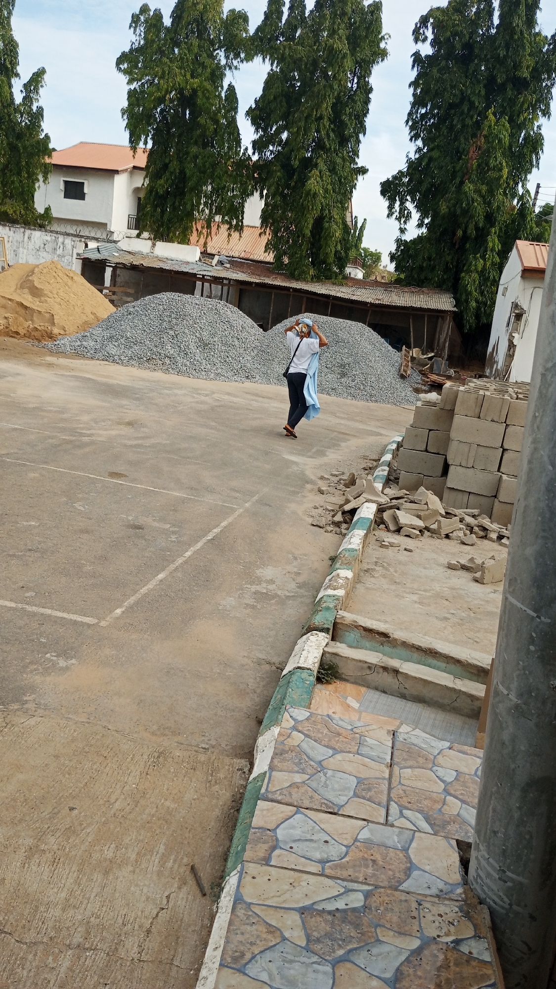 This outdoor image, taken in Lokoja, Nigeria, captures a scene that appears to be a construction site or material storage area. A person is walking away from the camera in the midground, dressed in a white t-shirt, dark trousers, and wearing a light blue fabric draped over their head and shoulders, which they are adjusting with both hands.
The ground beneath them and extending into the foreground is a large expanse of concrete or asphalt, marked with faint white lines, possibly remnants of a court or designated area. In the background, large piles of building materials are prominent: a light brown pile of sand to the left and several larger grey piles of gravel further back.
Beyond the construction materials, tall, slender green trees with dense foliage rise towards a bright, cloudy sky. Between the trees and above the material piles, a large white building with a distinct orange or reddish-brown tiled roof is visible, suggesting a residential or institutional structure. Smaller, more rustic buildings with corrugated metal roofs are also scattered behind the material piles.
To the right side of the image, a stack of grey concrete blocks is neatly arranged, next to a pile of broken concrete rubble. In the immediate foreground on the right, there's a decorative tiled area with steps leading downwards, featuring a pattern of varied earthy and grey tones. A thick, dark metal pole stands vertically on the far right. The overall lighting suggests it is daytime with clear weather. No text is visible in the image.