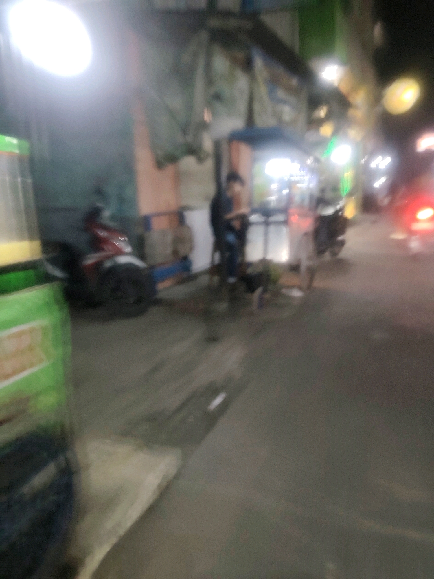 The image depicts a blurry street scene at night, likely in Mekarmukti, Indonesia, with several elements suggestive of a marketplace or food stall area. A bright, out-of-focus light source dominates the upper left quadrant. In the mid-ground, a person is standing near a red and white motorcycle, which is parked on the side of a paved area. Another person, also blurry, is visible in the background near some stalls or structures. A green container, possibly a dispenser or cooler, is in the foreground on the left. The ground appears wet, and there are reflections of lights on the surface. The overall impression is that of a busy, dimly lit outdoor commercial area with vehicles and people present.