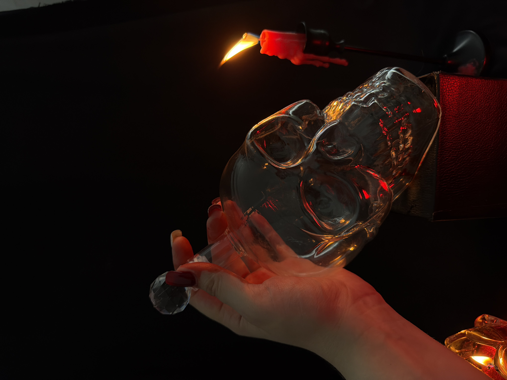 A hand with red nail polish is holding a glass skull decanter by its stopper. The decanter is placed on top of a dark red, textured surface, possibly a book. To the right, a red candle in a black candlestick burns, its flame casting a warm glow. Another candle, partially visible in the lower left corner, also emits light. The background is entirely black, creating a dramatic and mysterious atmosphere. The lighting is dim, with strong red and orange hues reflecting off the glass skull and the surfaces. The scene evokes a sense of gothic or occult aesthetics.