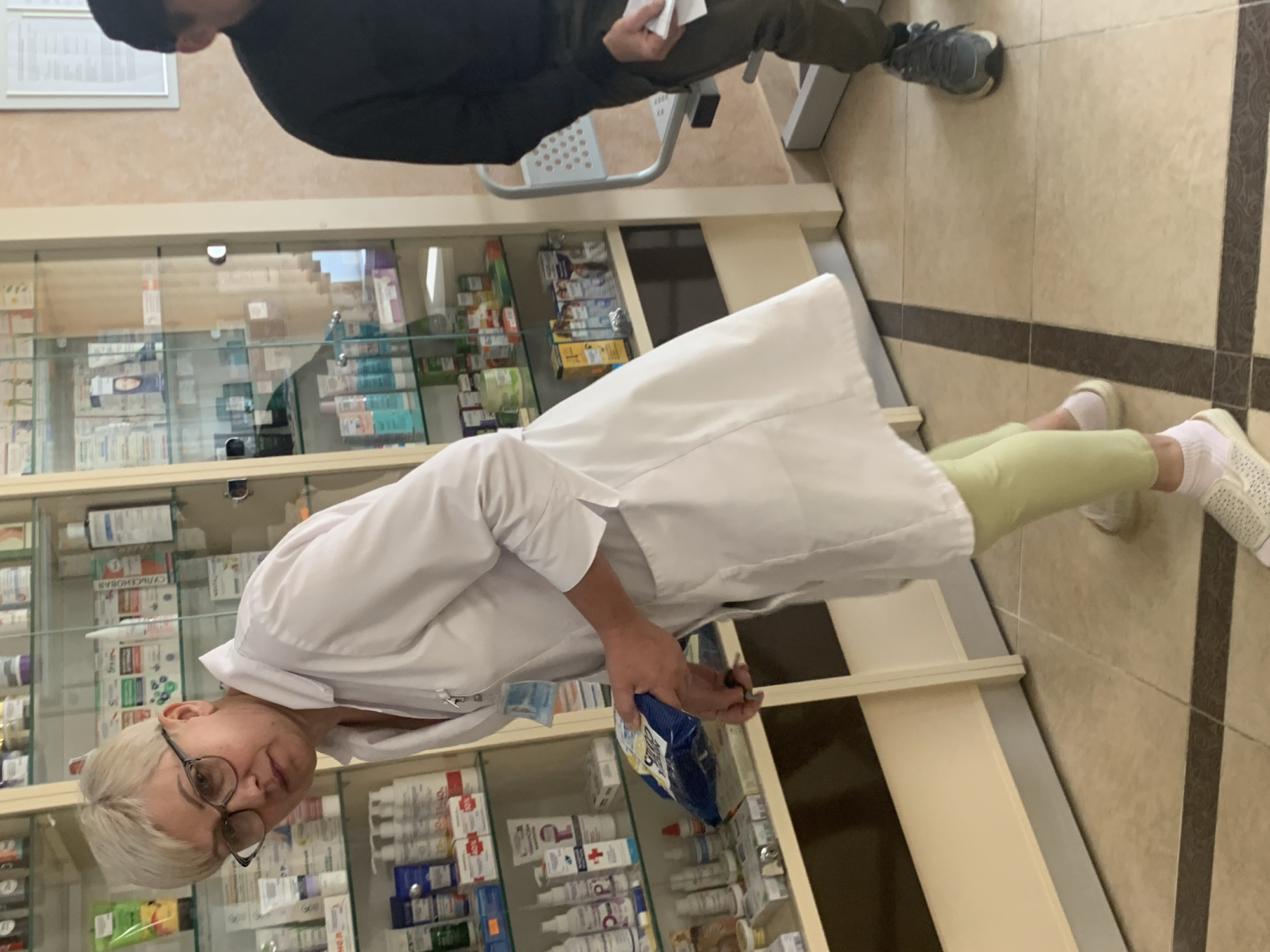 A blonde woman in a white lab coat and light green pants stands in a pharmacy in Kapyĺ, Belarus. She is wearing glasses and holding a blue package in her hands. The pharmacy has shelves stocked with various medications and health products. A man in a dark jacket and pants is seated to the right of the woman.