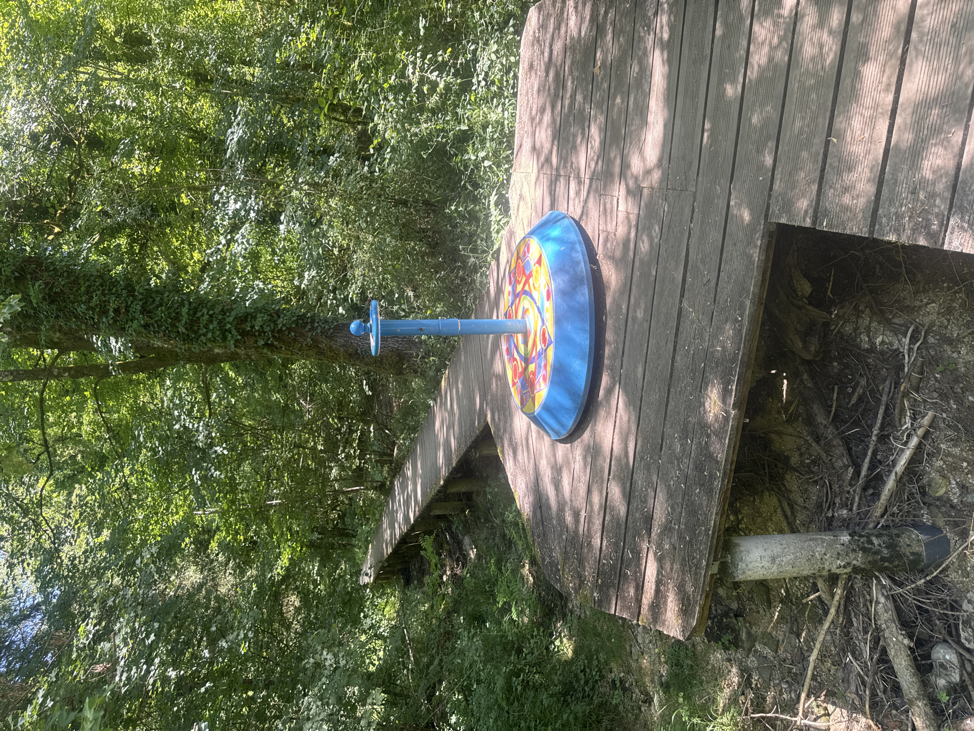 A wooden boardwalk winds through a sun-dappled forest in Ravilloles, France. In the foreground, a circular, blue, outdoor game or play structure with colorful geometric patterns sits atop a wooden platform. The structure has a central blue pole with a knob at the top. The boardwalk is elevated, supported by wooden posts, and appears to be constructed from weathered planks. The surrounding environment is dense with green foliage, including trees and undergrowth, with sunlight filtering through the leaves. A small, dry creek bed or ditch is visible beneath a section of the boardwalk. The scene suggests a recreational area within a natural setting.